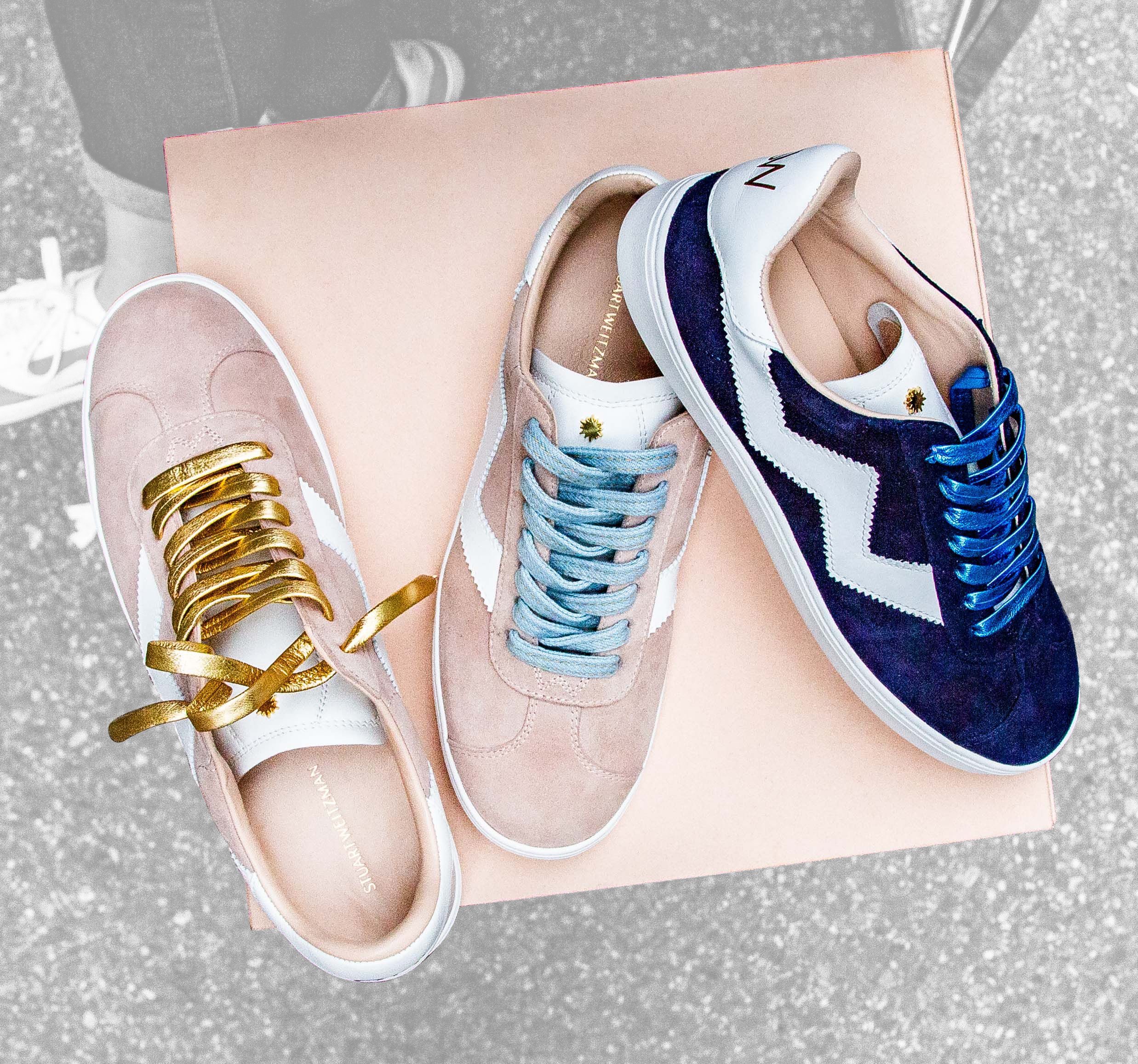 Three suede sneakers—two light pink and one navy—sit on a pale pink box with colored laces.