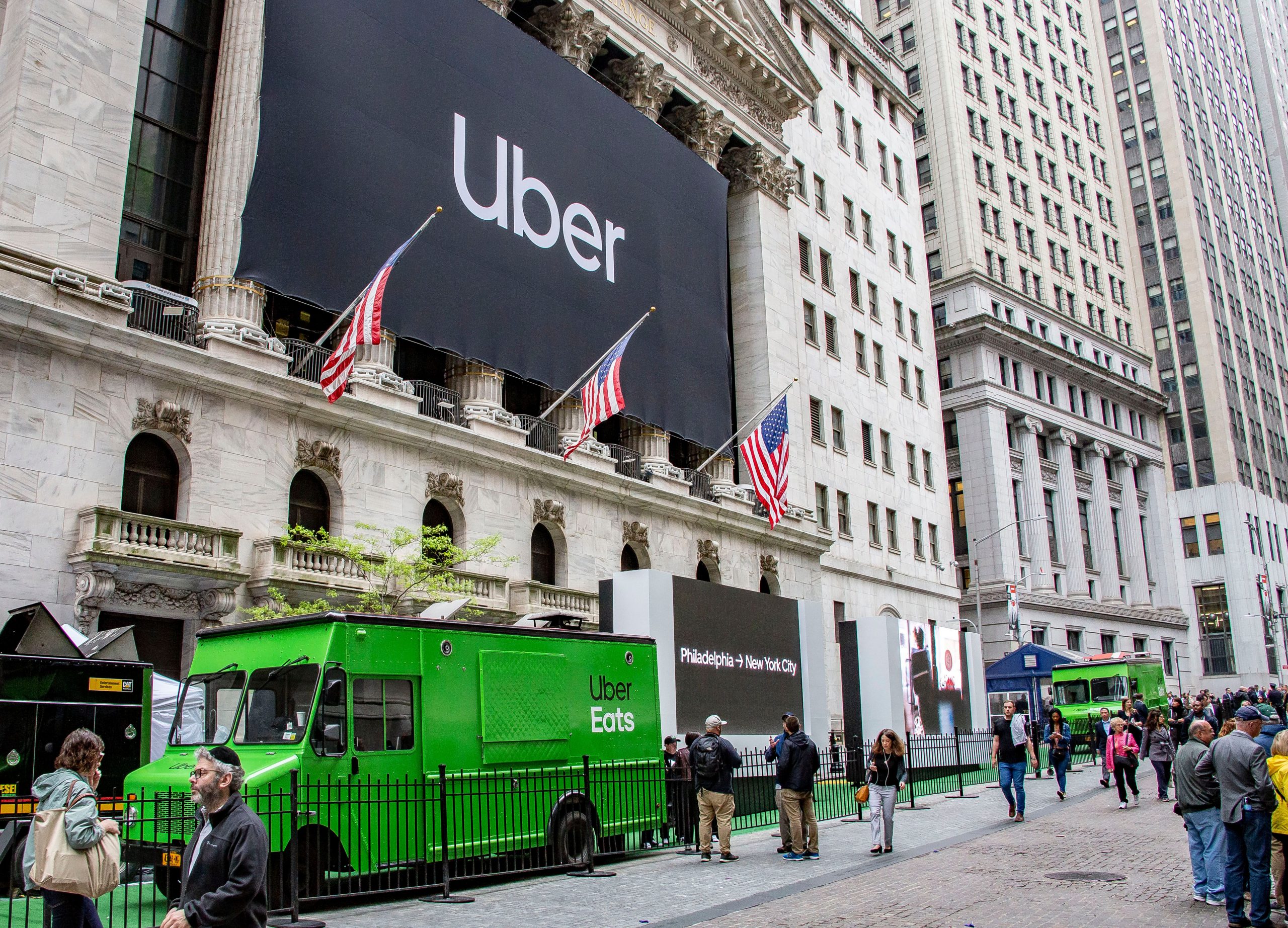 Two green Uber Eats trucks parked outside the New York Stock Exchange with an Uber banner and flags.