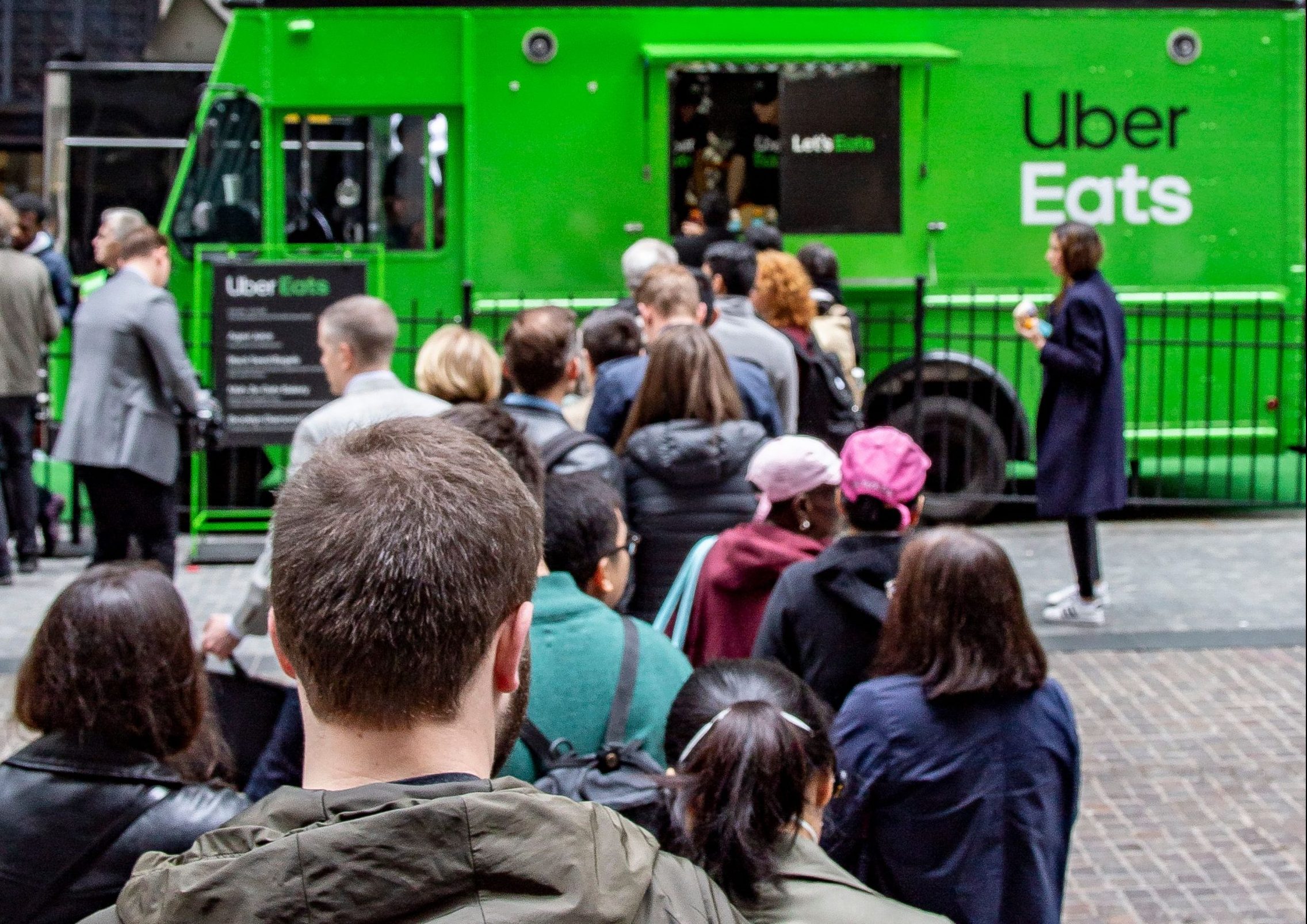People wait in line at a green Uber Eats food truck parked on a busy city street.