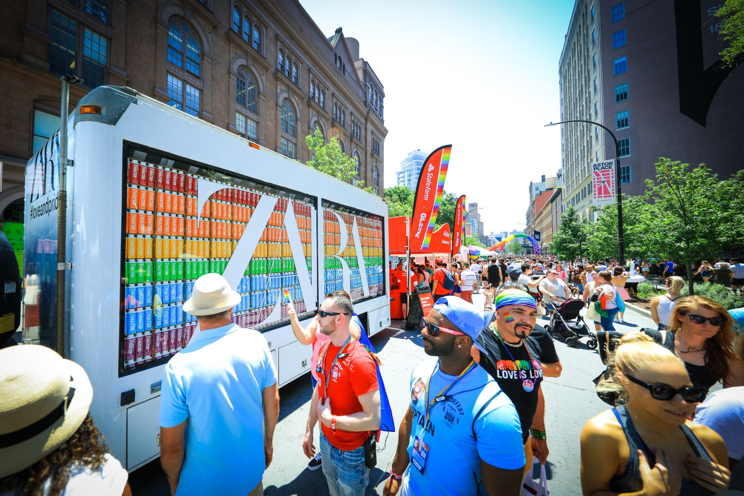 Street parade with people, rainbow flags, and a truck with cans spelling ZAP; crowds celebrate.