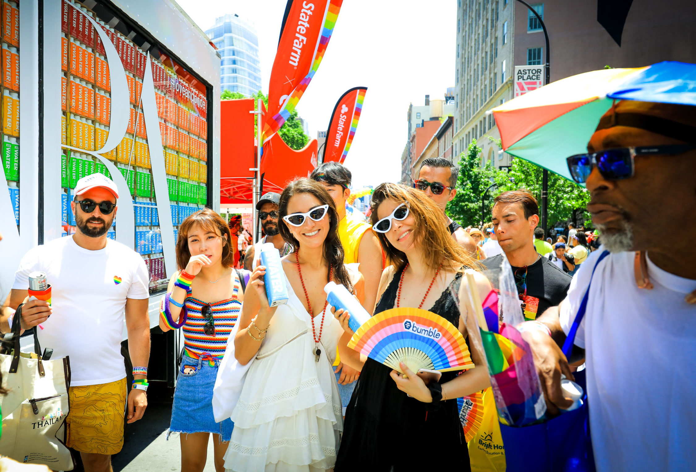 People celebrate at an outdoor Pride event with rainbow accessories and city buildings behind.