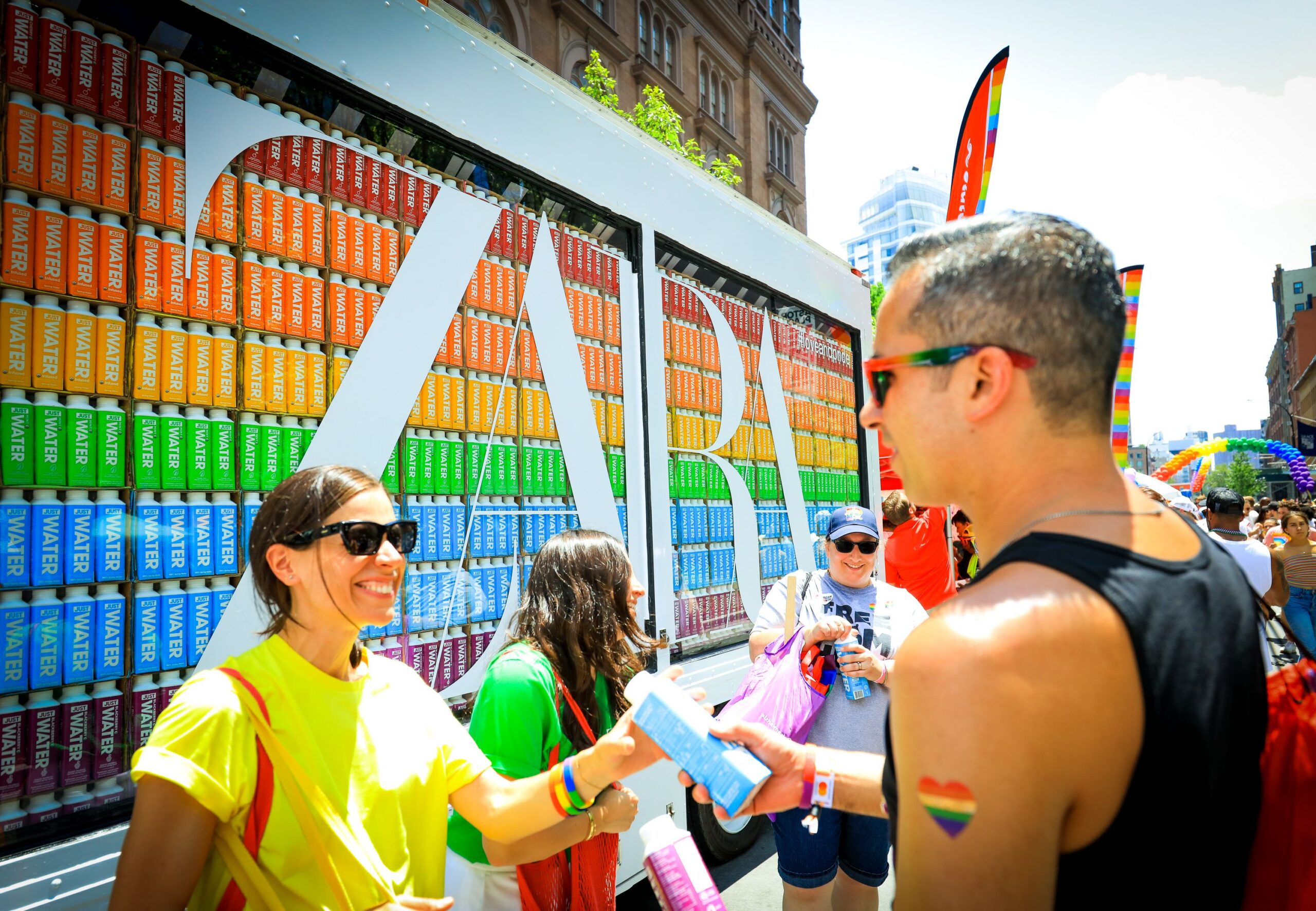 People in colorful clothes and rainbow stickers gather outside a ZARA display of rainbow boxes.