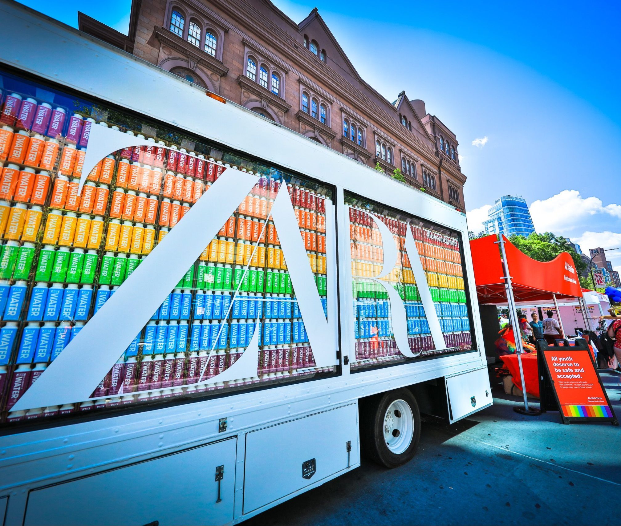 A truck with “ZARU” branding and stacked cans parked by a red booth with people in a city square.