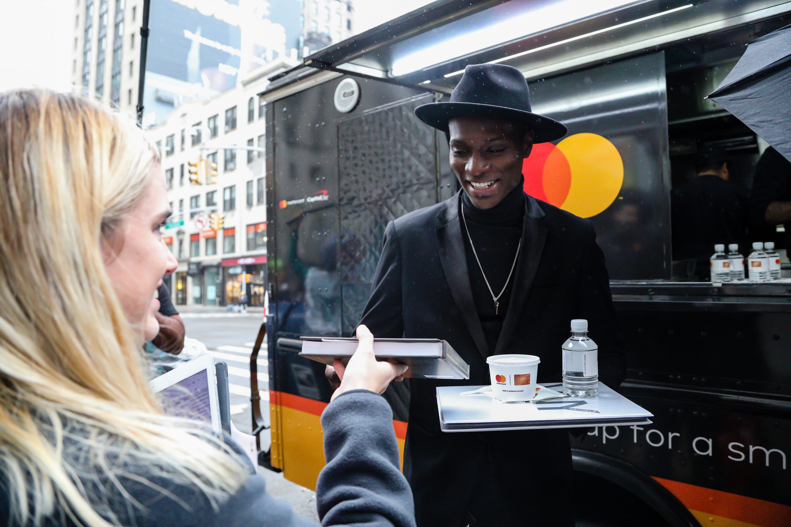 Man in black hands food tray with drink and notebook to woman at Mastercard vehicle on city street.