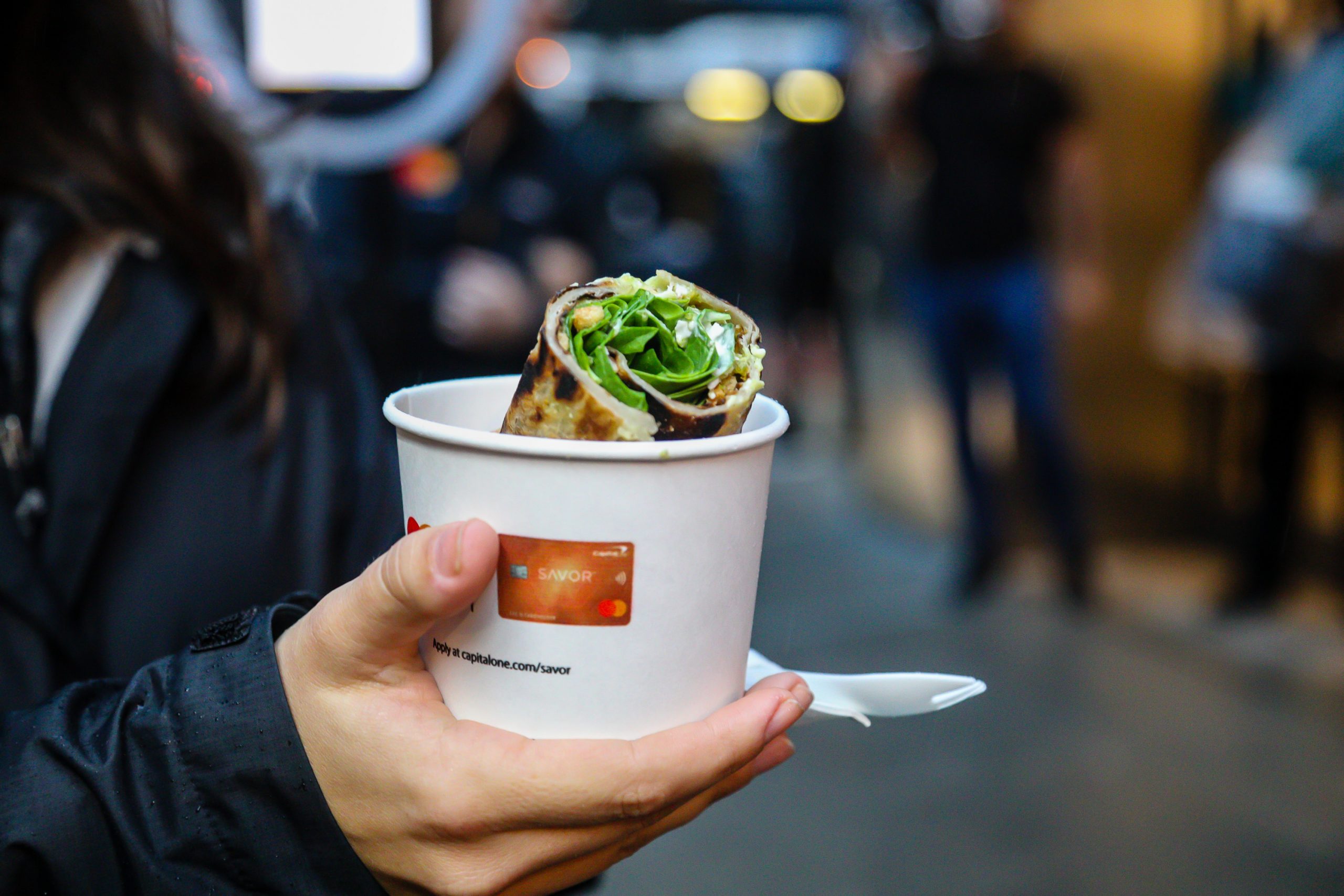 Person holds a white cup with a rolled wrap and greens, plastic spoon attached, at a market.