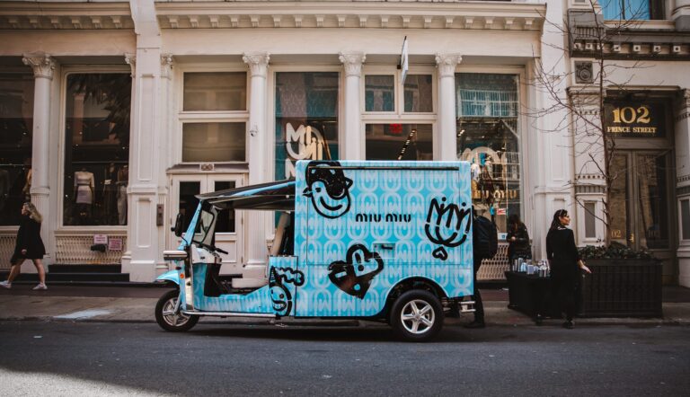 Blue tuk-tuk with cartoon designs parked by a white building; person stands nearby at a table.