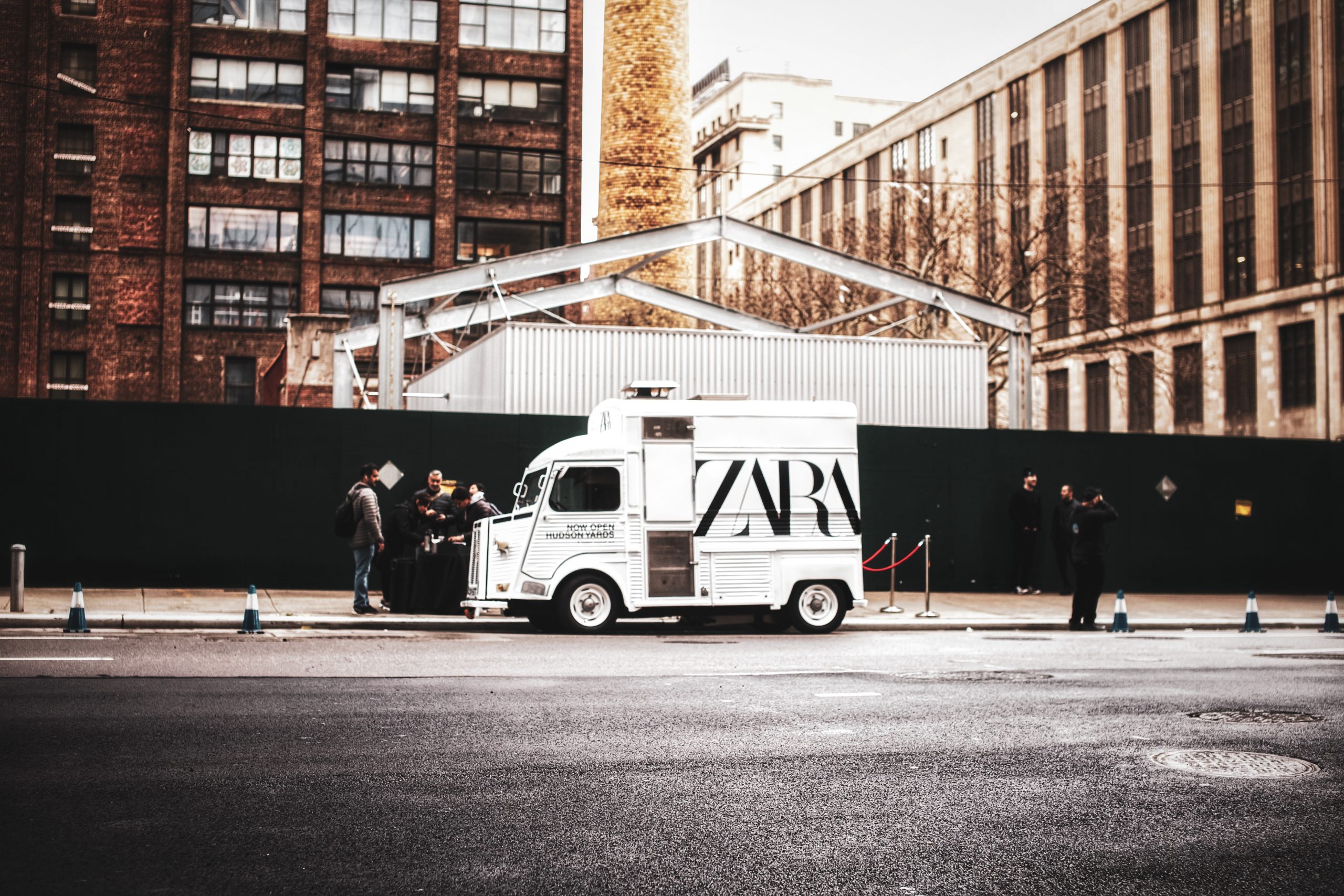 A white Zara vehicle is parked on a city street with several people and brick buildings behind it.