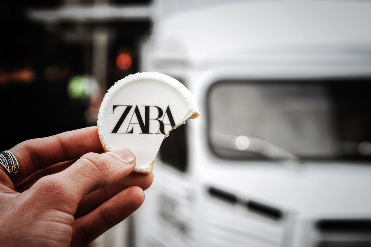 Hand holding a bitten white cookie with the black ZARA logo; blurred vehicle in background.
