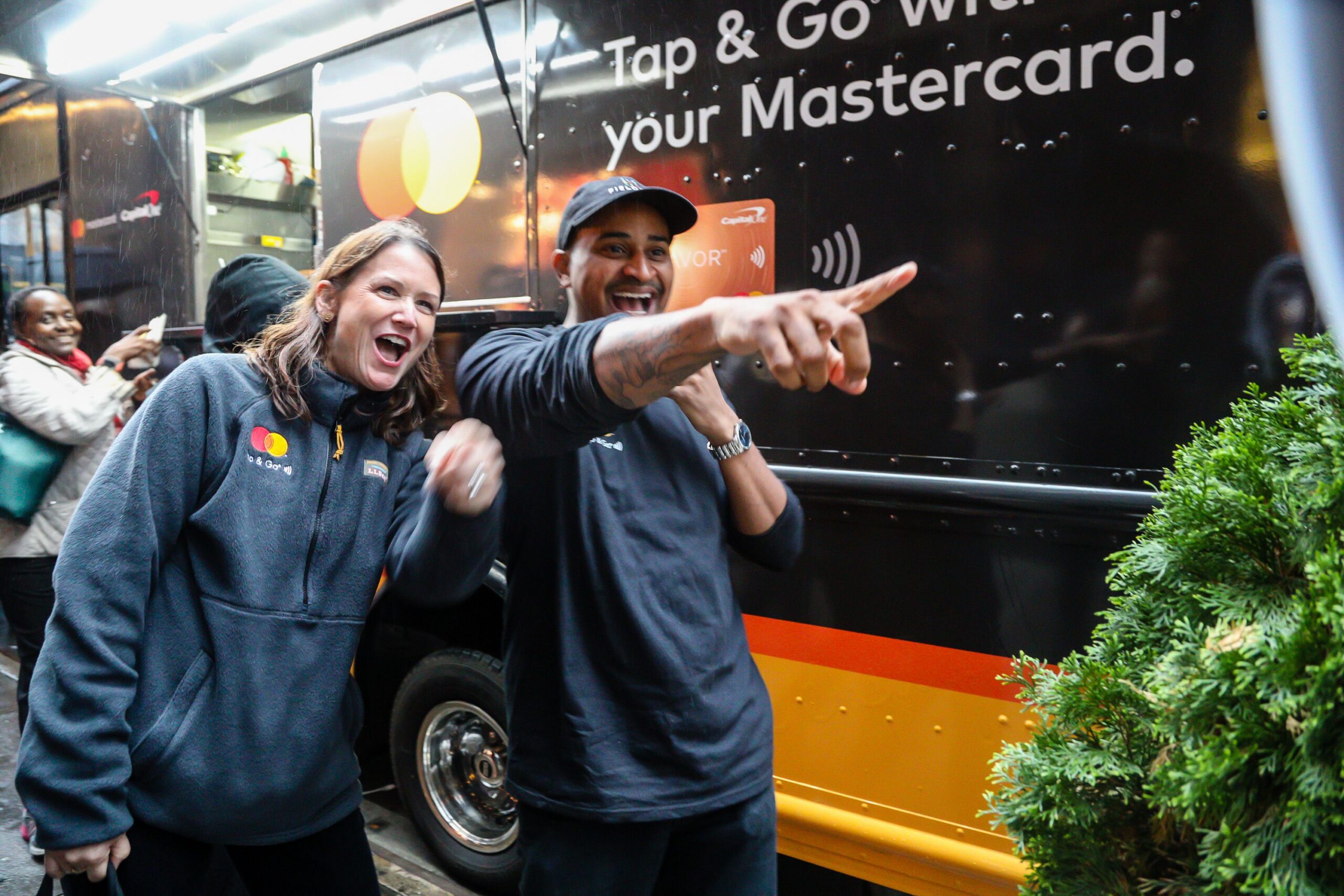Two people smile and point in front of a Mastercard vehicle; others and greenery are behind them.