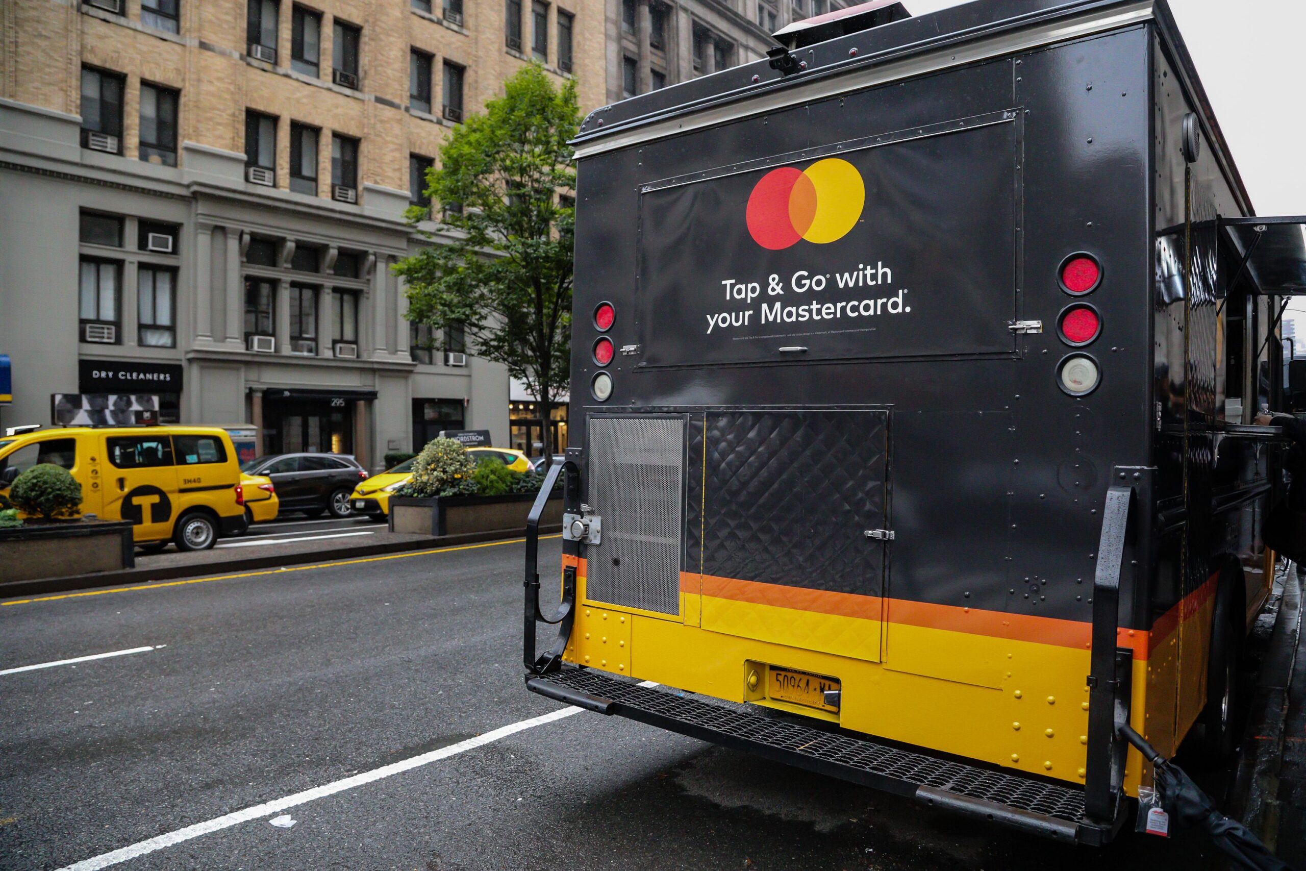 Black Mastercard promo vehicle parked on city street, near yellow taxis and buildings.