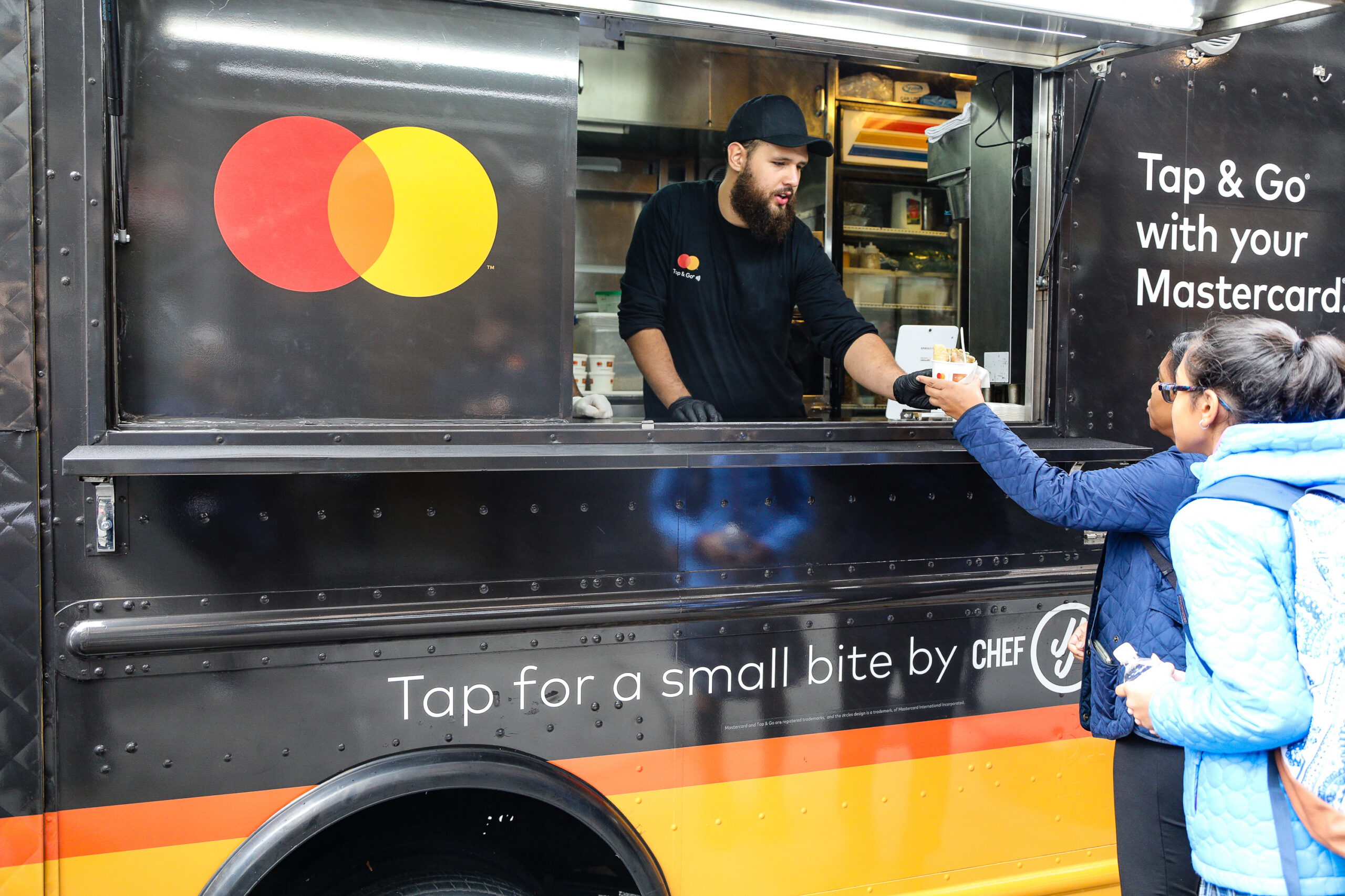 Man in food truck hands drink to woman; Mastercard logos and Tap & Go messages on vehicle.