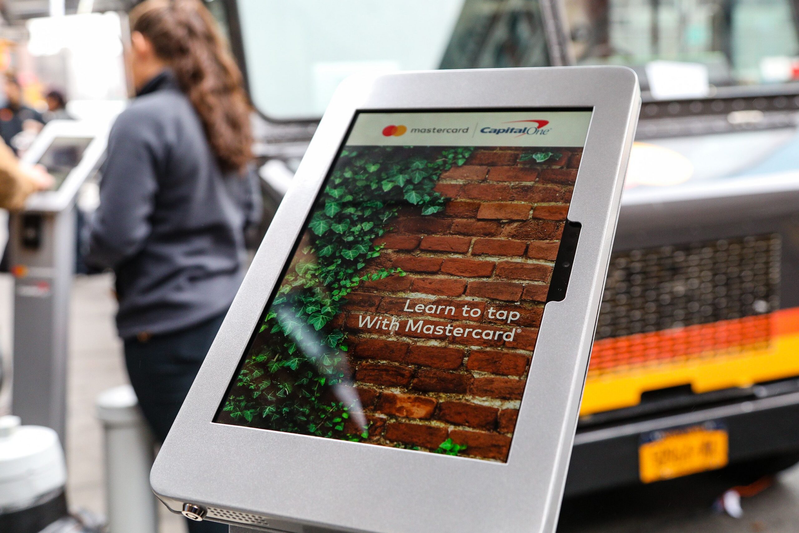 Touchscreen kiosk with "Learn to tap With Mastercard" and logos; people, promo vehicle behind.
