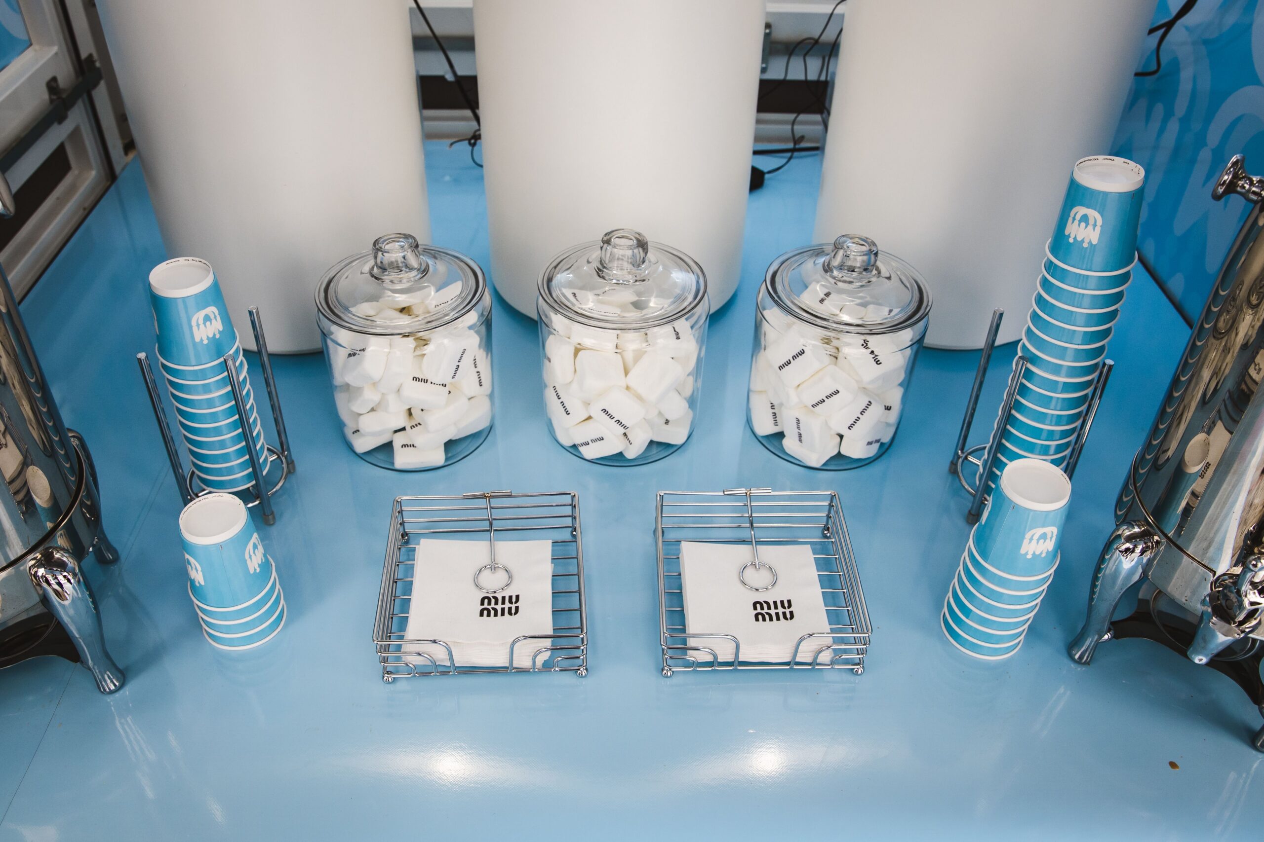 Glass jars of sugar cubes, striped cups, and miu miu napkins on a light blue countertop.