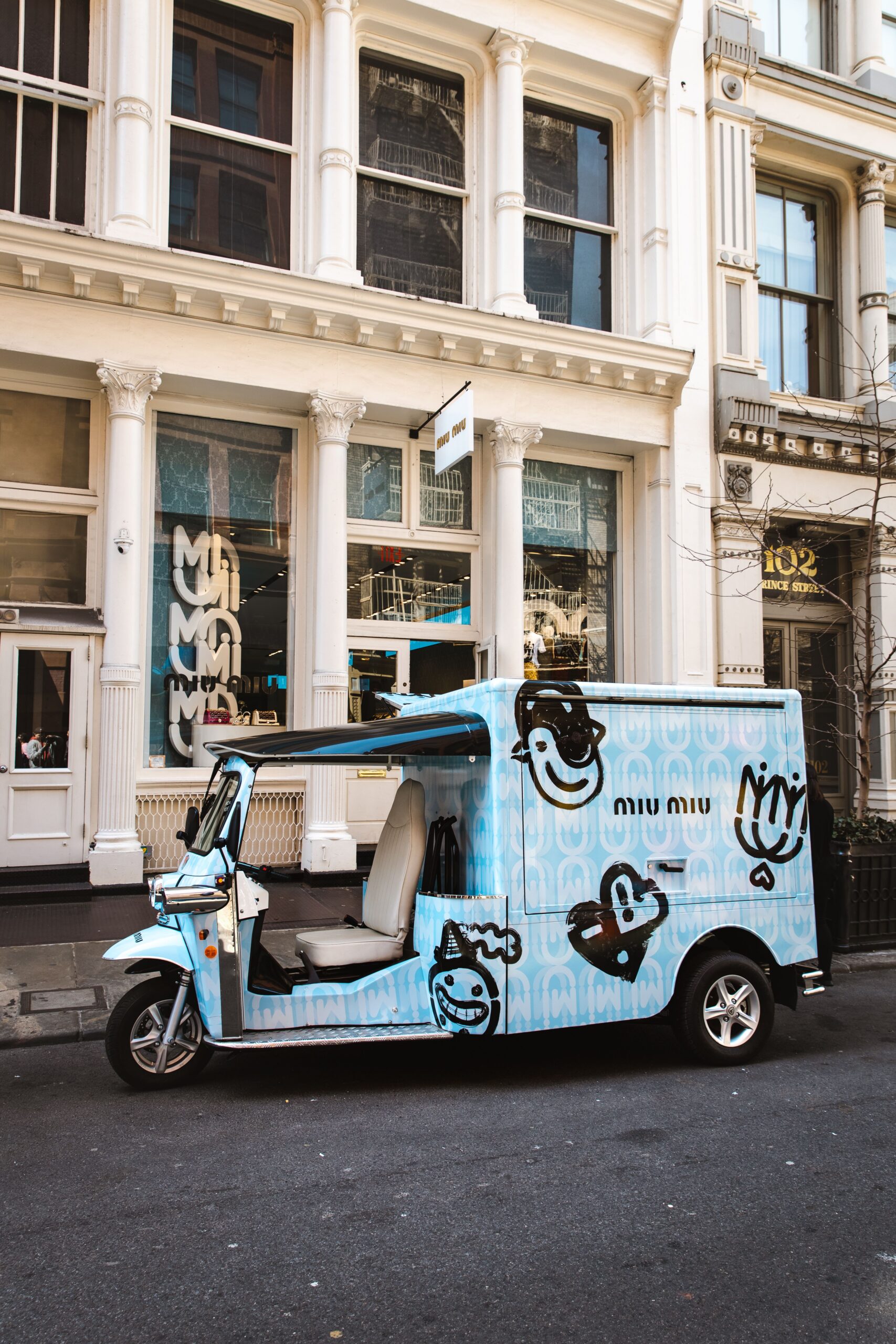 Light blue three-wheeled delivery vehicle with cartoon faces parked by a white building.