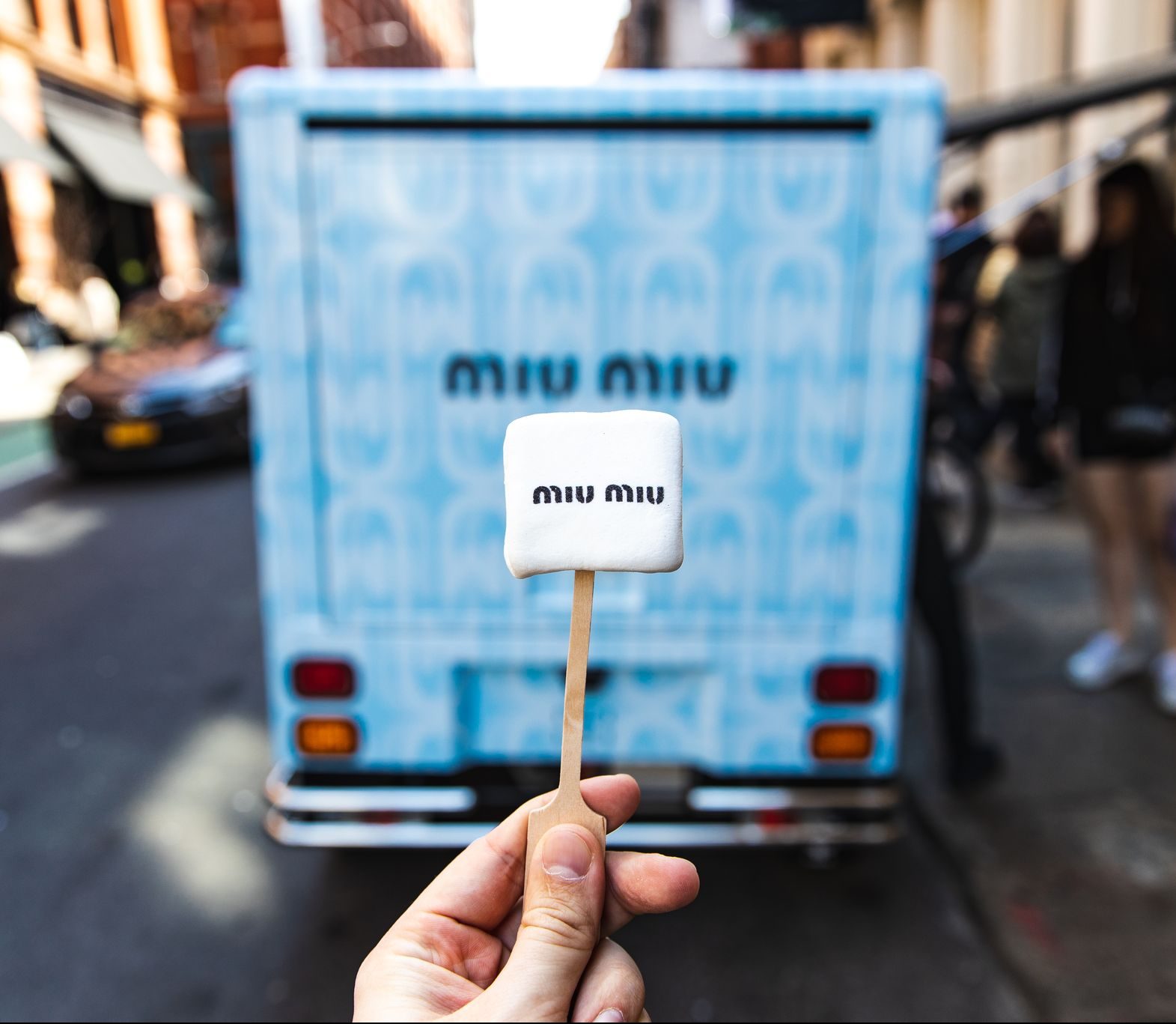 Hand holds marshmallow with miu miu logo in front of a blue truck showing the same logo.