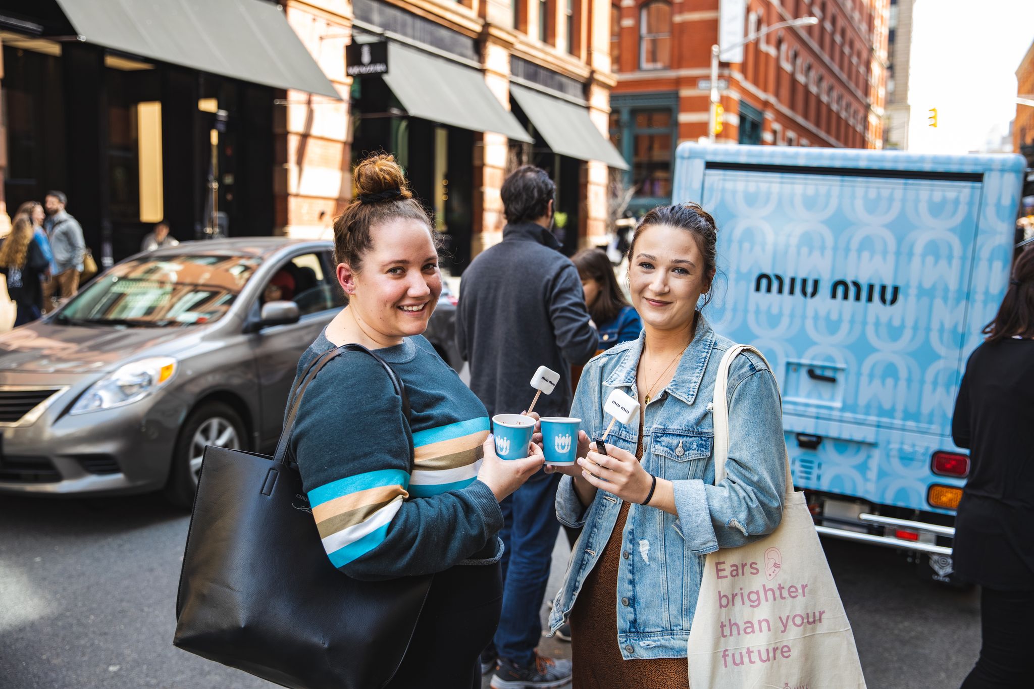 Two women smiling with ice cream on a city street, blue ice cream truck and cars behind them.