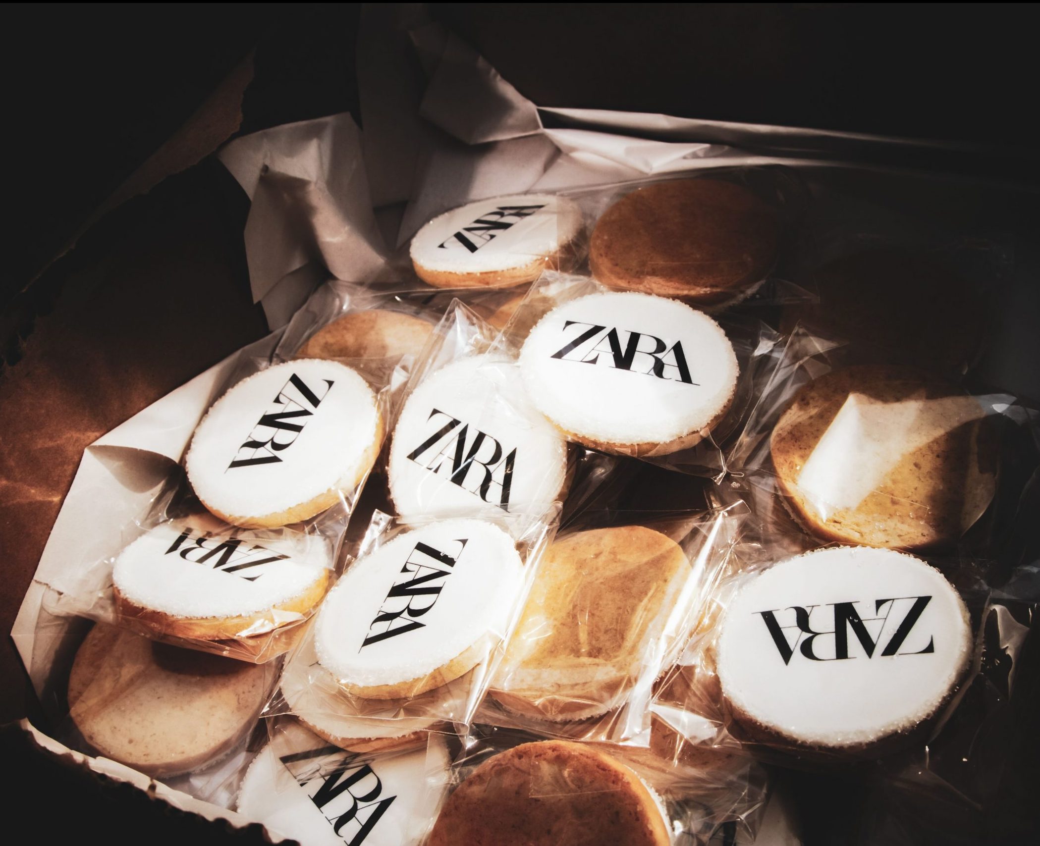 Box of round cookies, some with ZARA logo on white icing, wrapped; plain unwrapped cookies included.