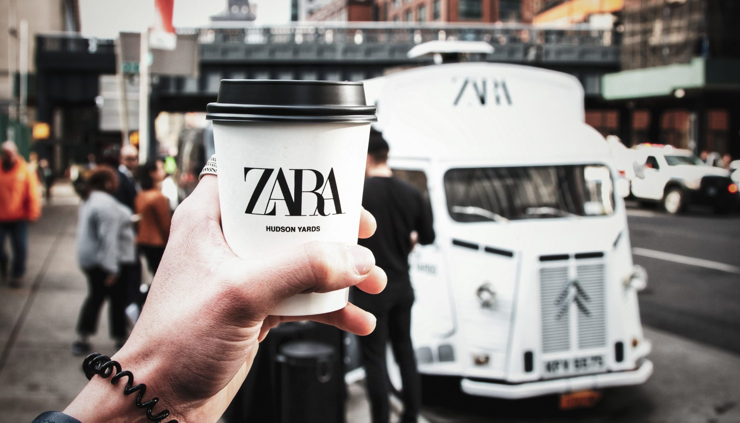 Hand holding a white ZARA coffee cup in front of a food truck on a busy city street.