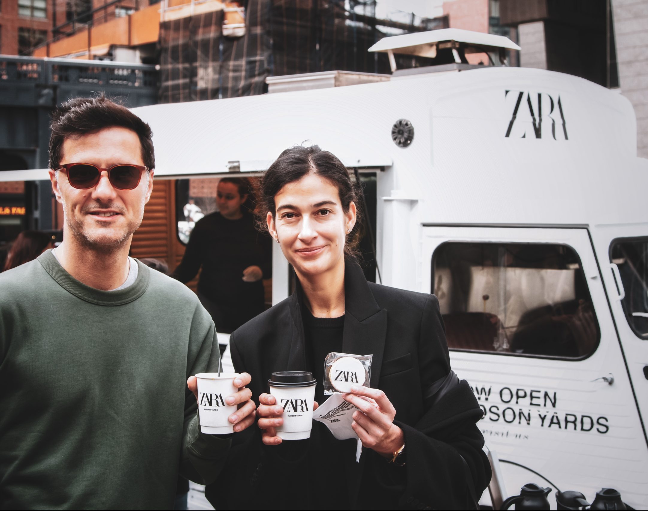 Two people with coffee stand by a white Zara vehicle, modern city buildings behind them.