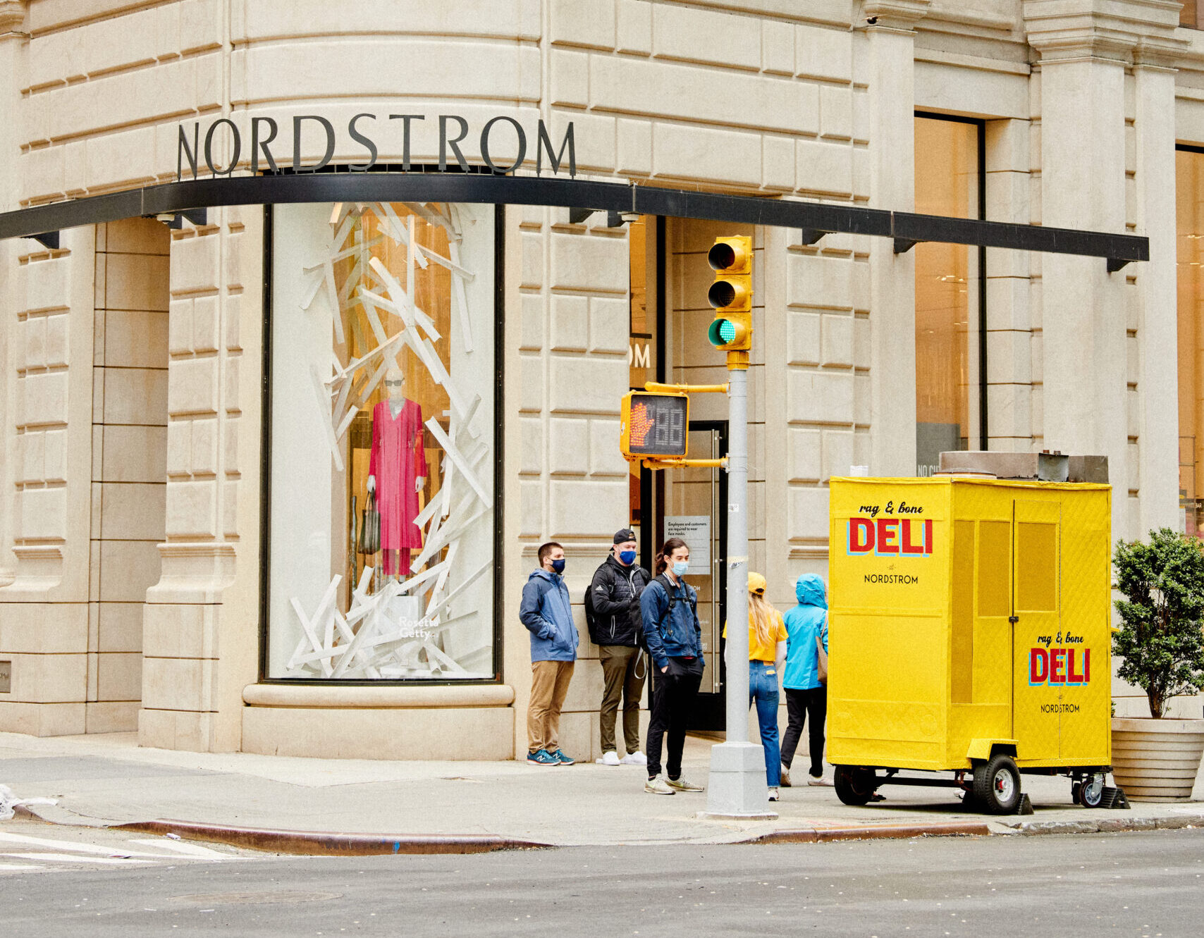 Four people at a street corner by Nordstrom with white displays and a yellow hot dog cart nearby.