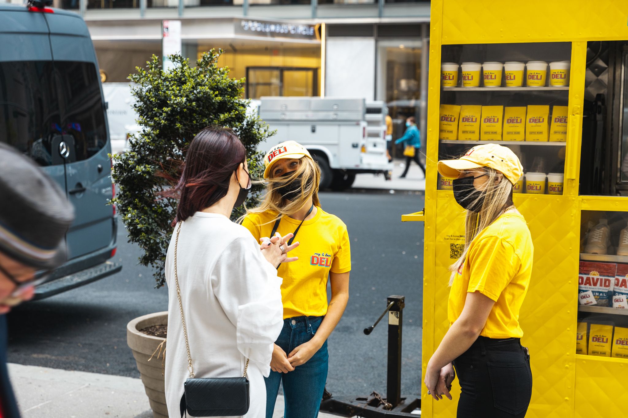 Two women in yellow by a food cart talk to a woman in white; all wear black masks on city street.