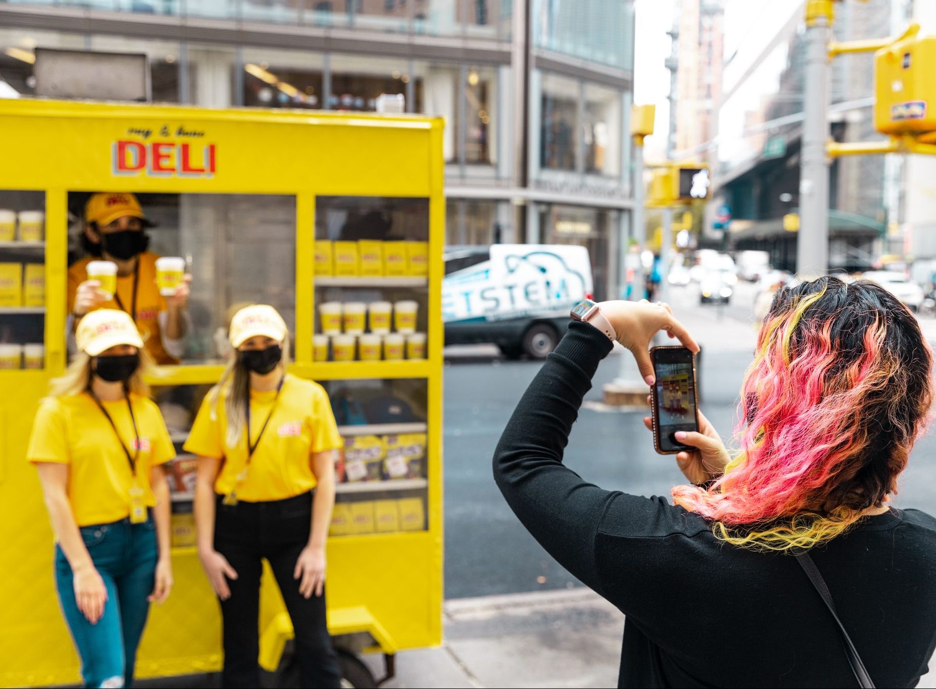 Two women in yellow shirts and hats by a deli cart, holding cups and wearing face masks.