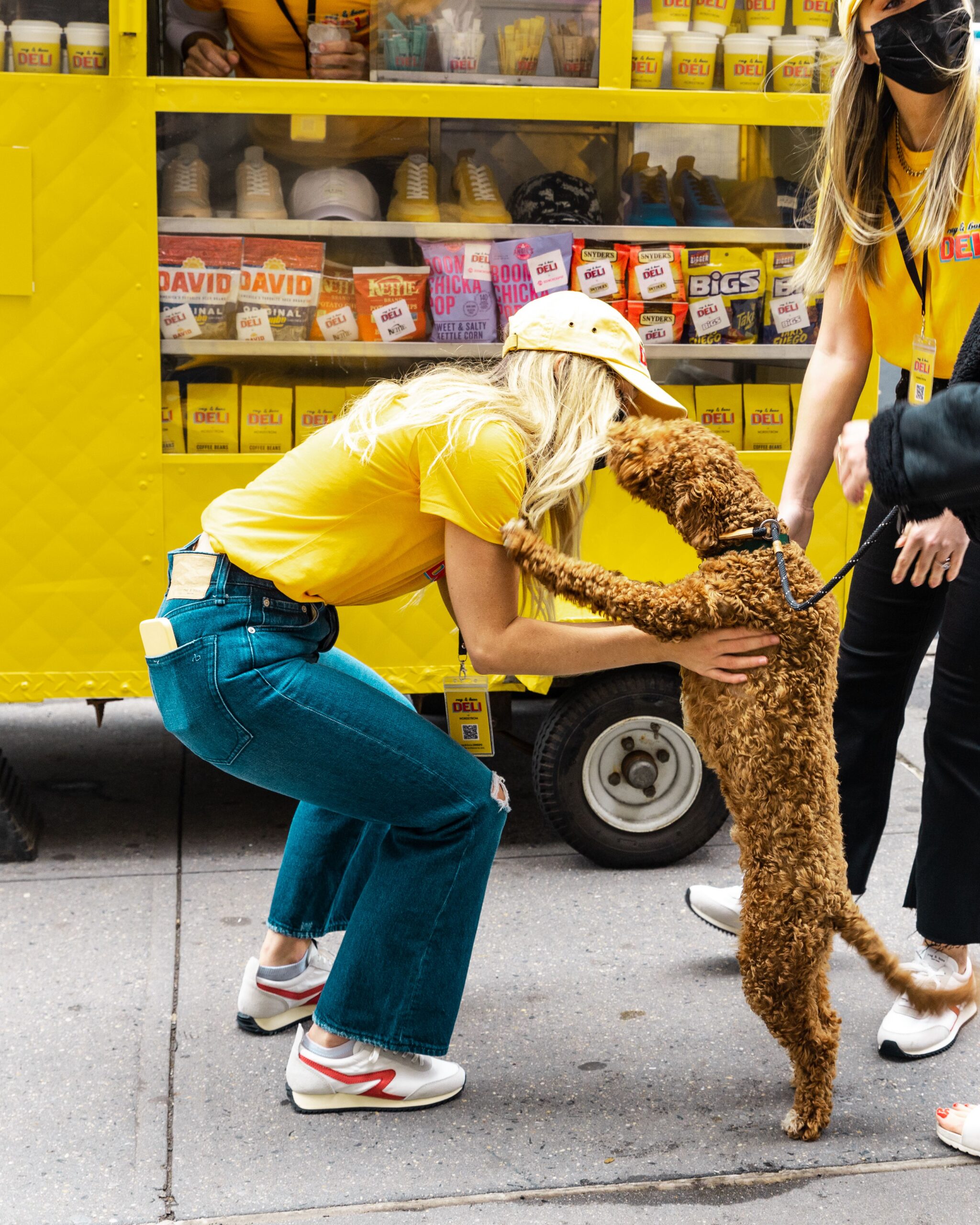 Woman in yellow hugs a standing brown dog as another holds its leash near a yellow food cart.