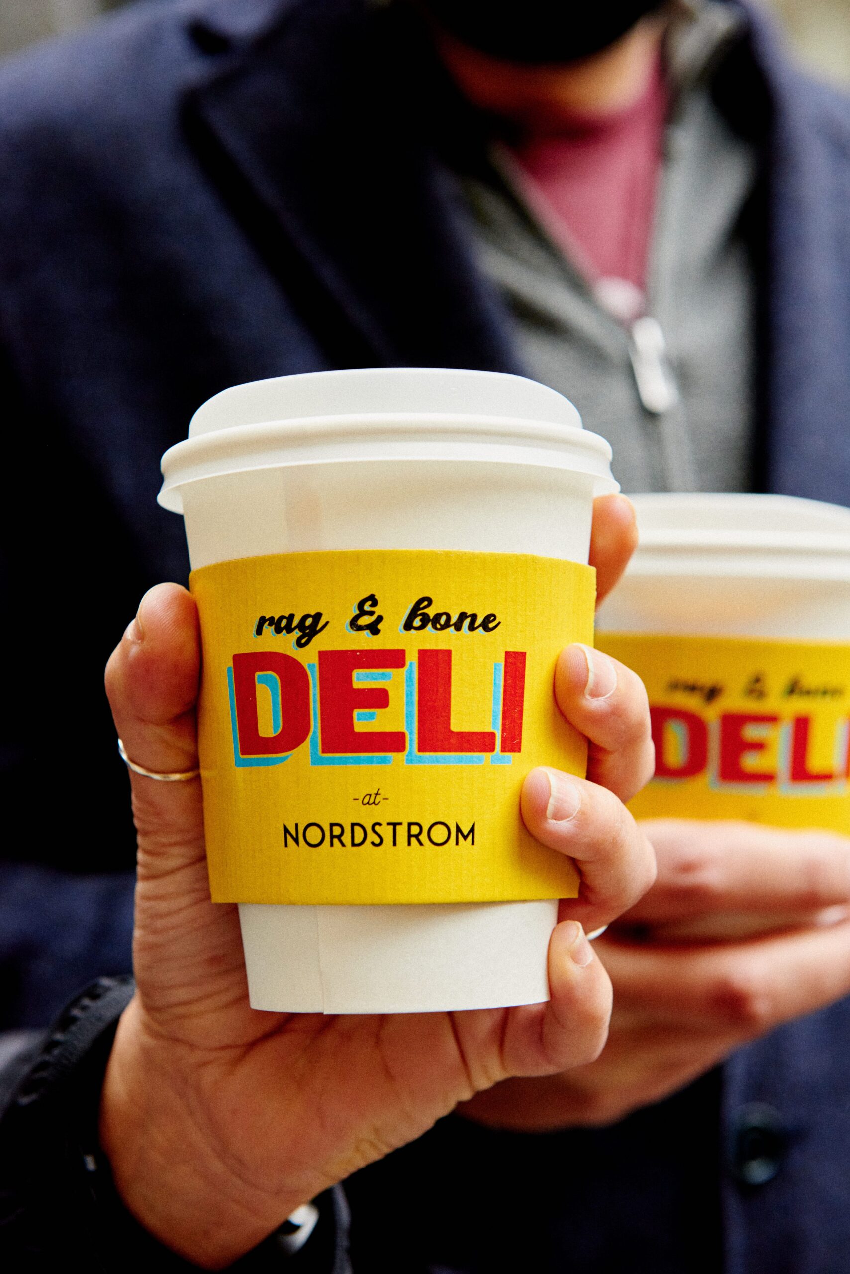 Hand holding a white coffee cup with yellow rag & bone DELI sleeve; another cup in background.