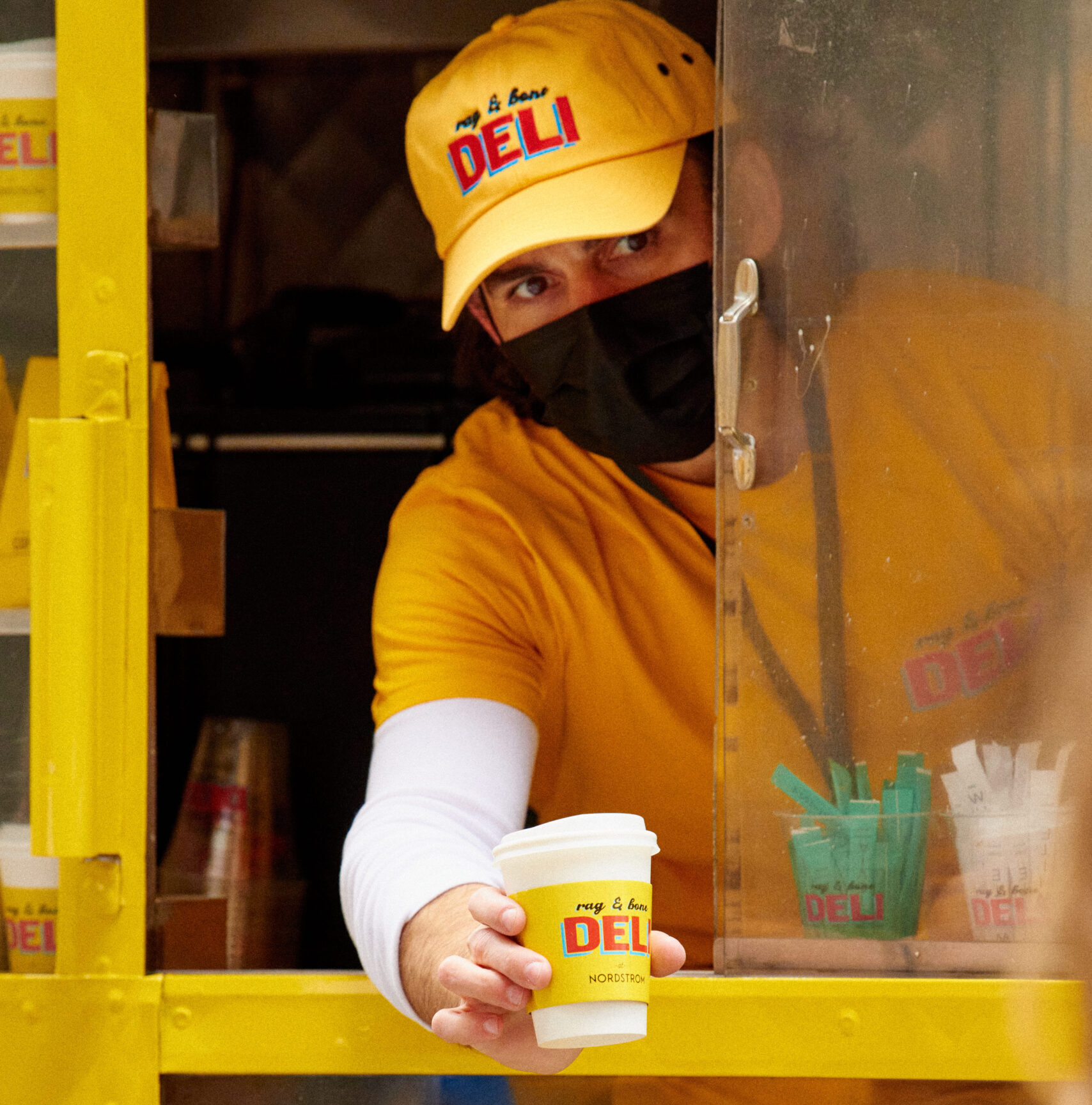 Person in yellow DELI cap and shirt hands coffee cup through yellow food truck window.