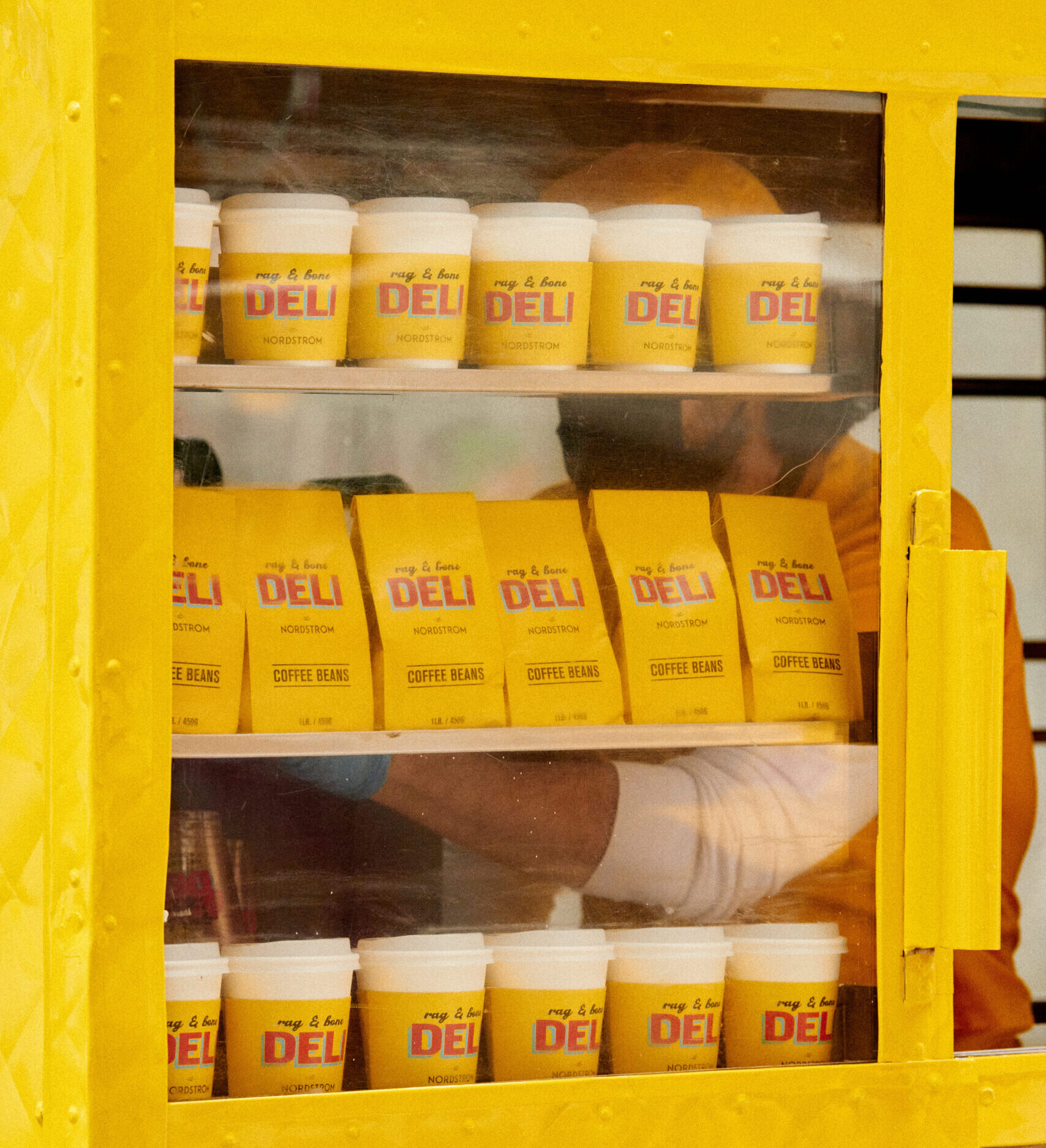 Shelves with yellow and white cups, "DELI" coffee bags, and a person in yellow behind the glass.