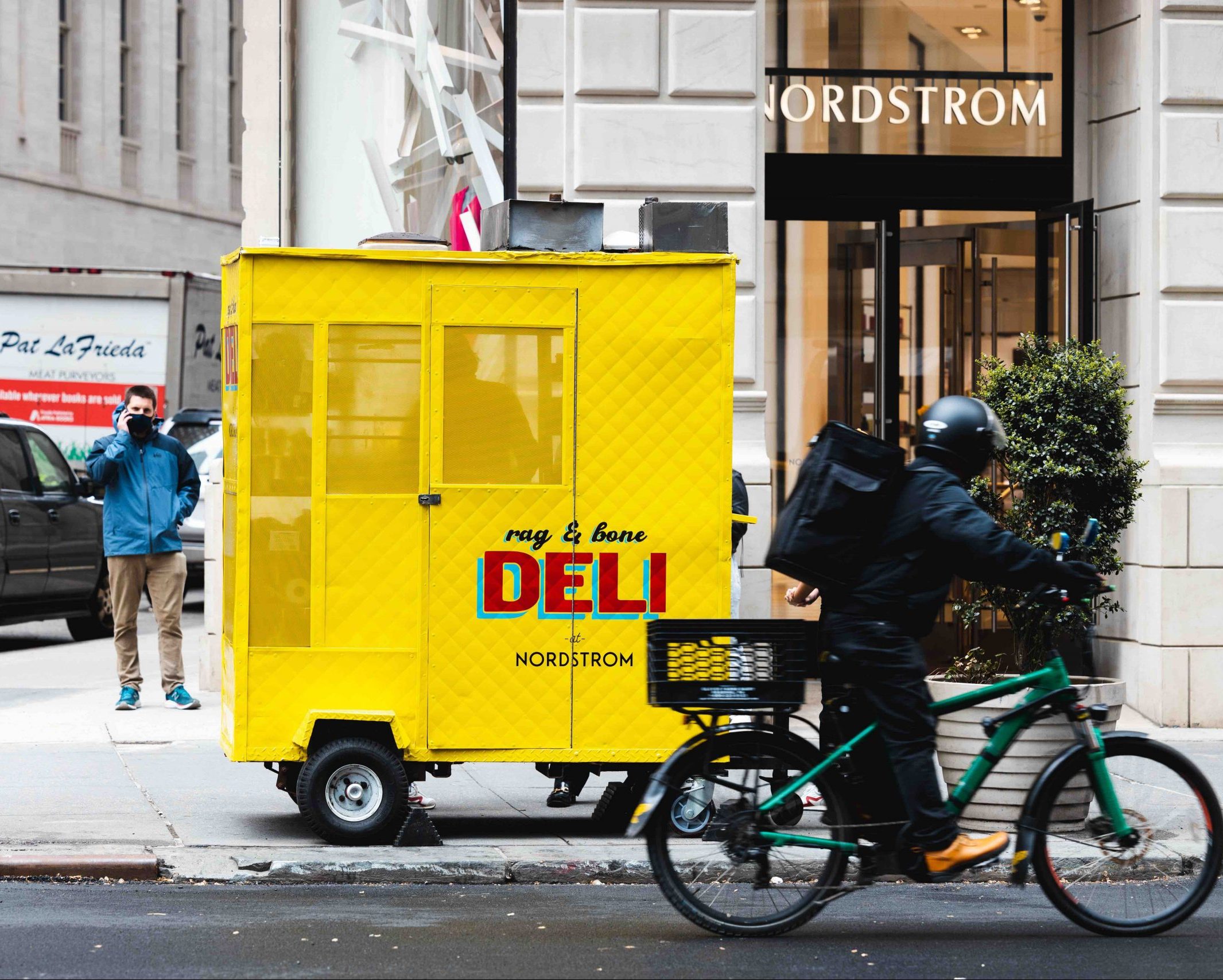 Yellow food cart marked rag & bone DELI NORDSTROM by Nordstrom; cyclist and person on phone nearby.