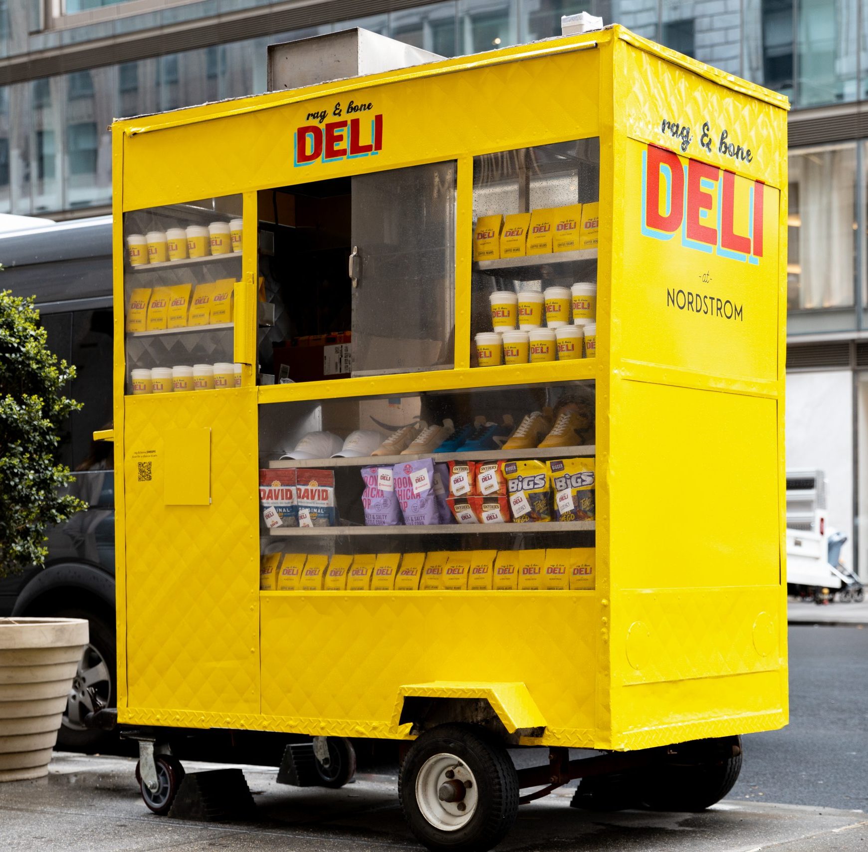 Yellow deli cart with "DELI" and "Nordstrom" signs, stocked with snacks, parked by a building.
