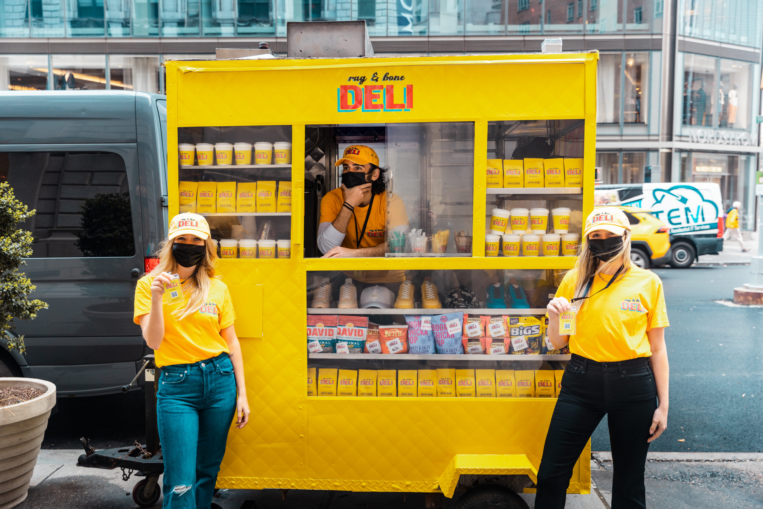 Two people in yellow deli uniforms hold drinks outside a yellow food cart; one person is inside.