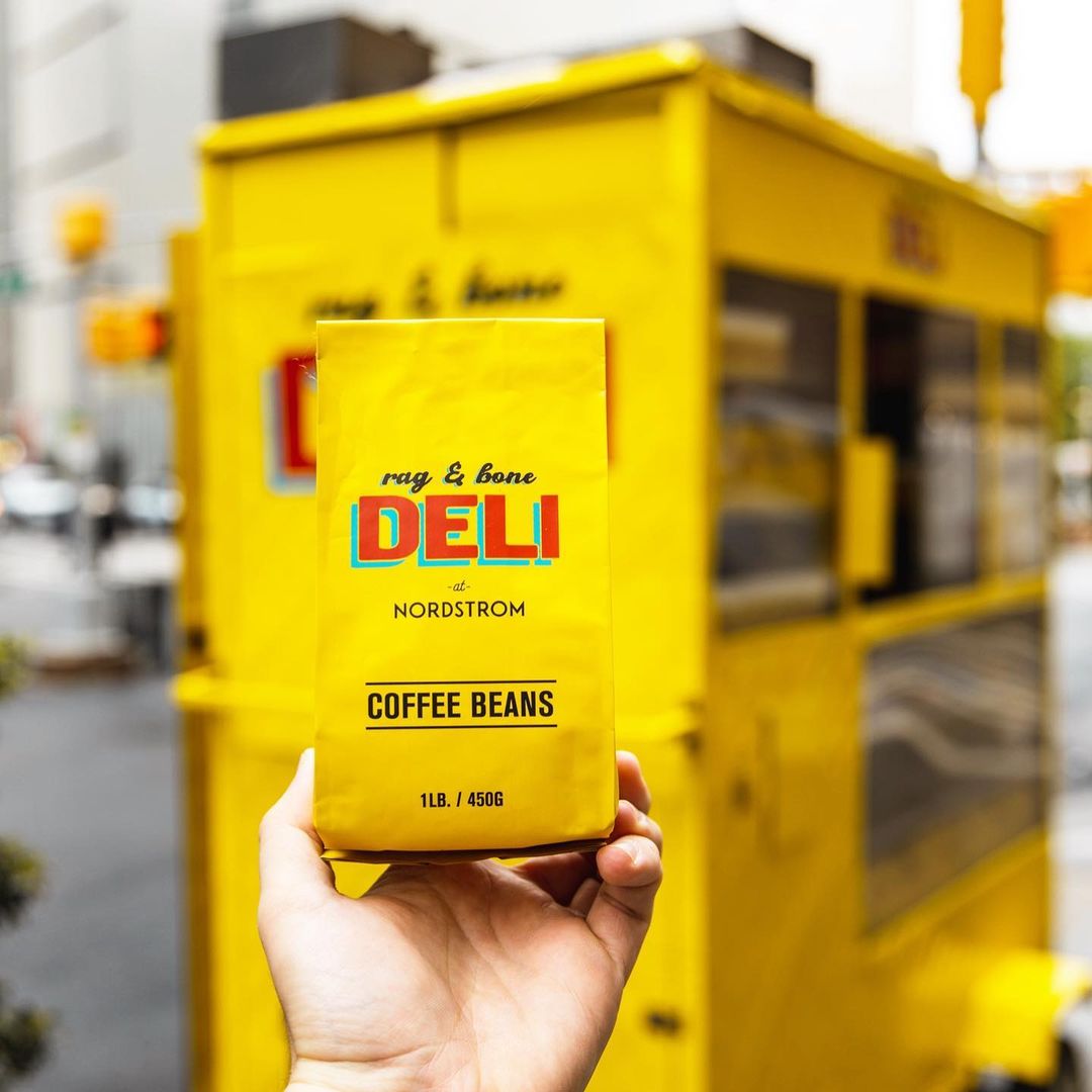 Hand holds a yellow rag & bone DELI at NORDSTROM coffee bag in front of a yellow food cart.