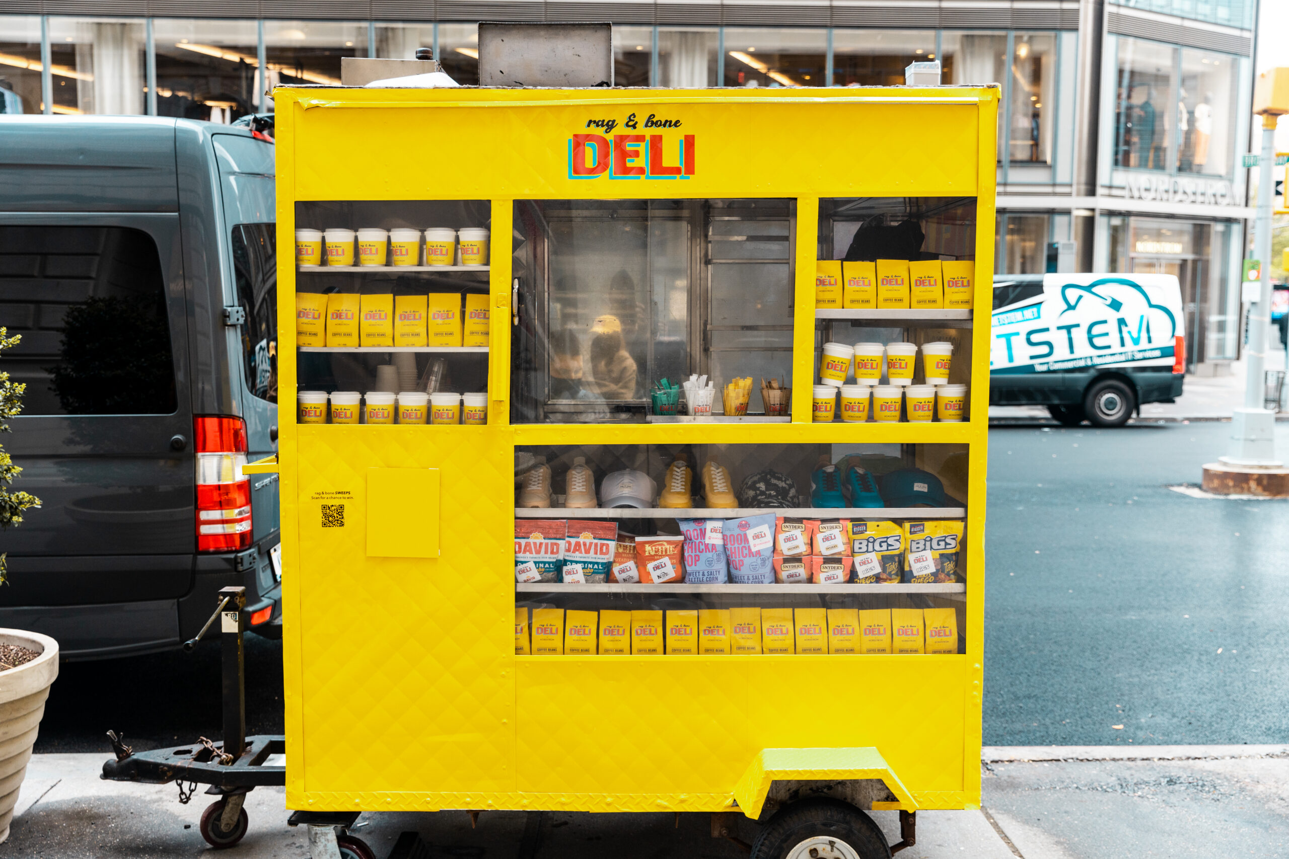 Yellow deli food cart on city street with snacks in window; buildings and a van behind.