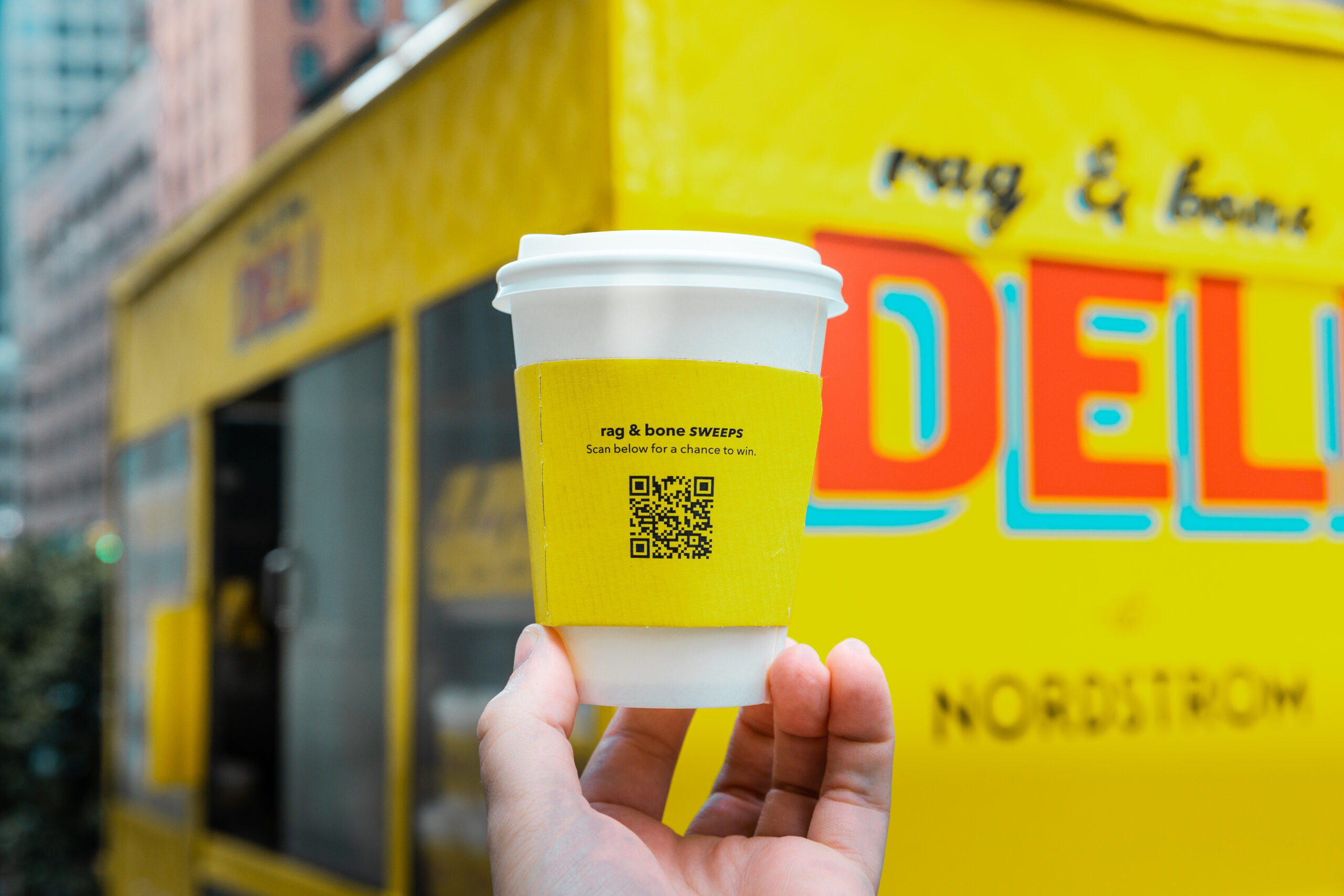 Hand holding a white coffee cup with yellow QR sleeve in front of a yellow food truck marked DELI.