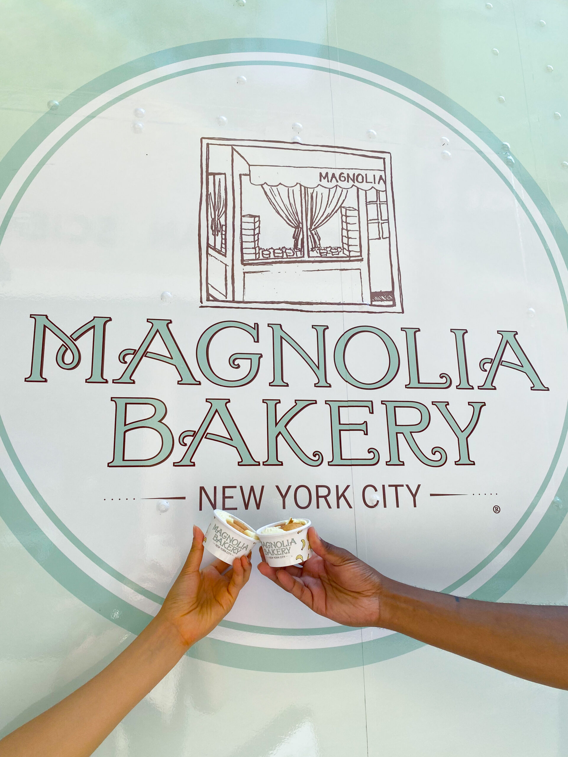 Hands hold banana pudding cups before a Magnolia Bakery logo and truck on a light blue background.