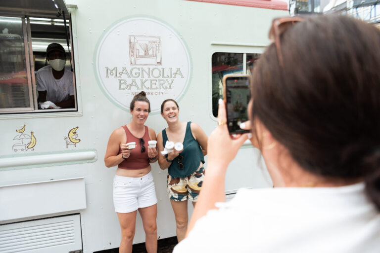 Two women holding desserts pose by the Magnolia Bakery truck as someone takes their photo.