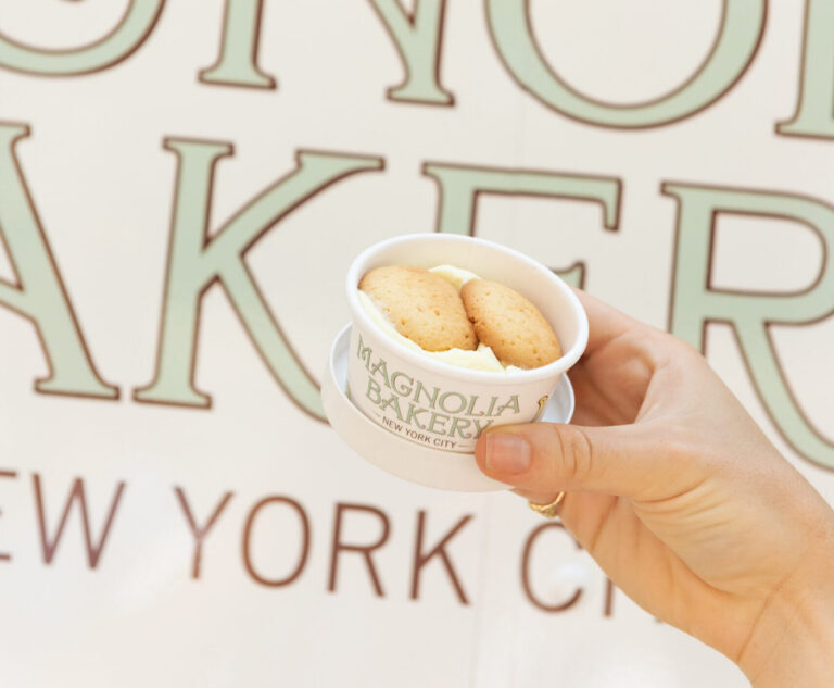 Hand holds Magnolia Bakery banana pudding with vanilla wafers; bakery truck blurred in background.