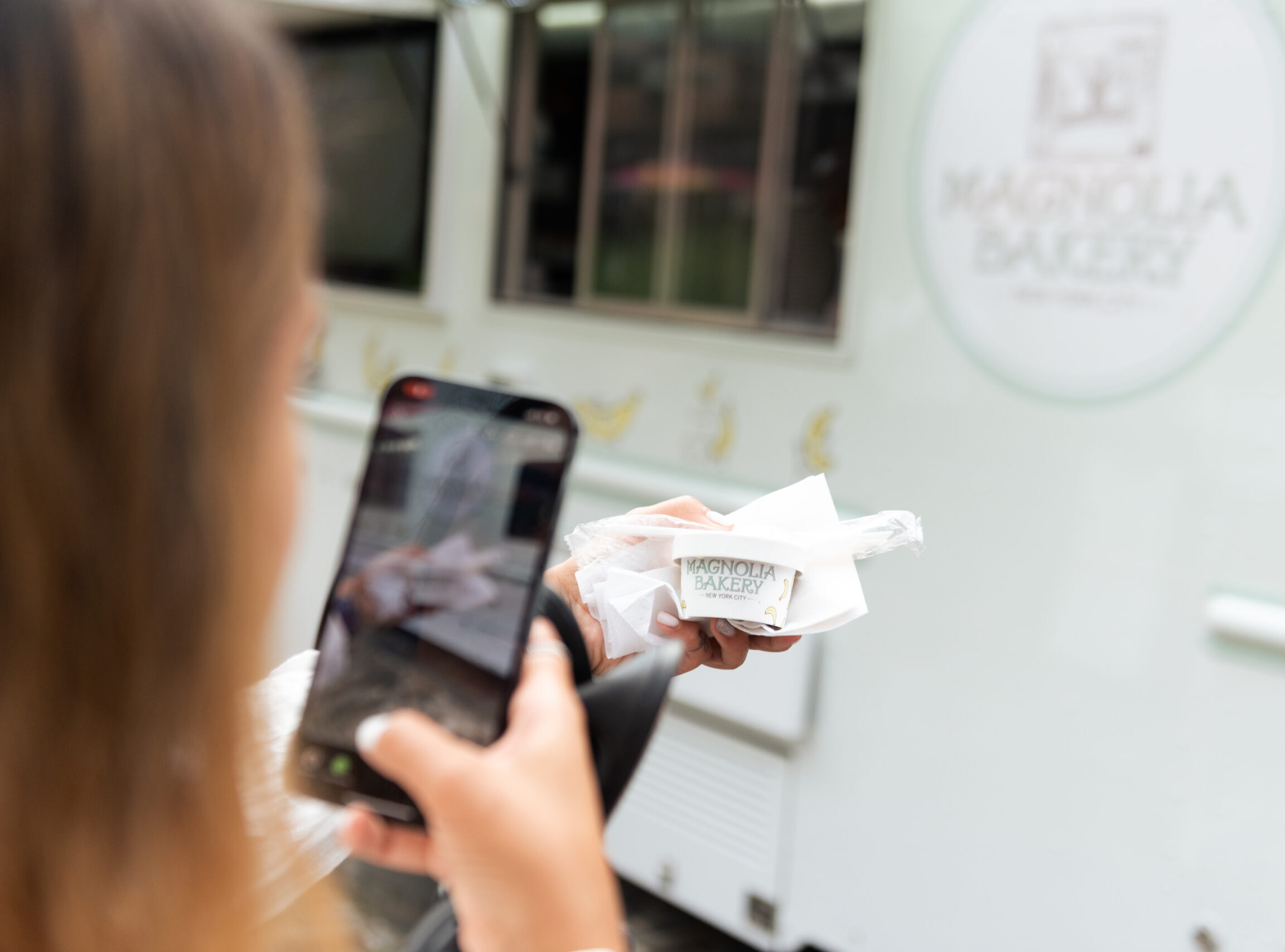 Person taking a phone photo of a Magnolia Bakery dessert cup in front of the bakery truck.