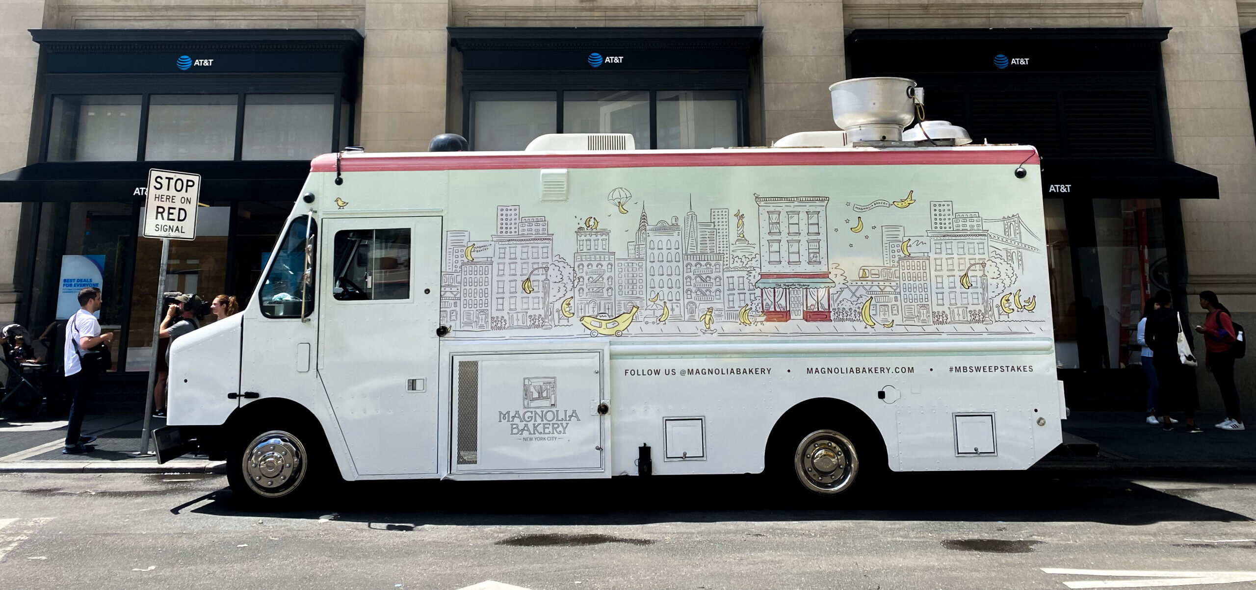 Magnolia Bakery truck with cityscape and banana art parked on street near people and stop sign.