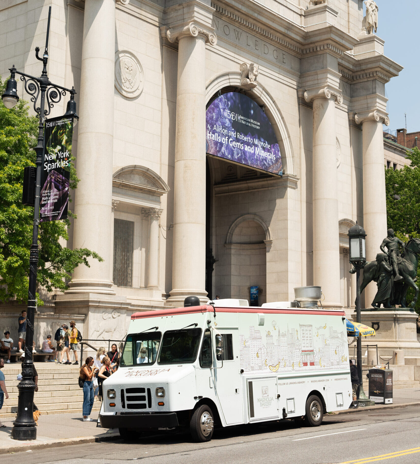 Magnolia Bakery truck parked outside a stone museum with columns; people walk nearby.