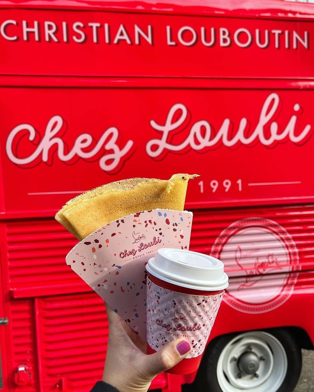 Hand holding a crepe and pink-sleeved coffee in front of the red Chez Loubi food truck.