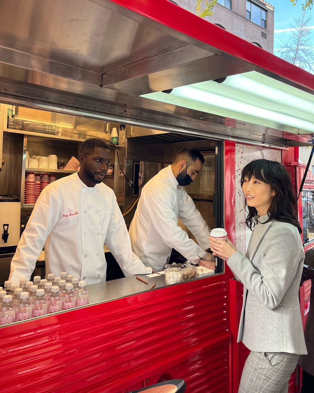 Woman in gray suit with coffee talks to two food truck workers at red pop-up with pastries.