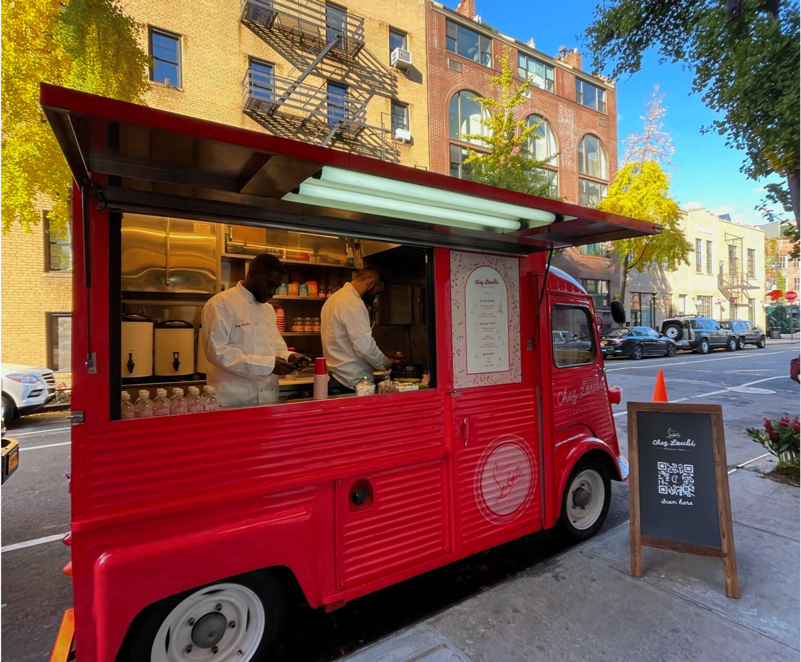 Two people cook in a red food truck on a city street; menu and QR code are on the side.