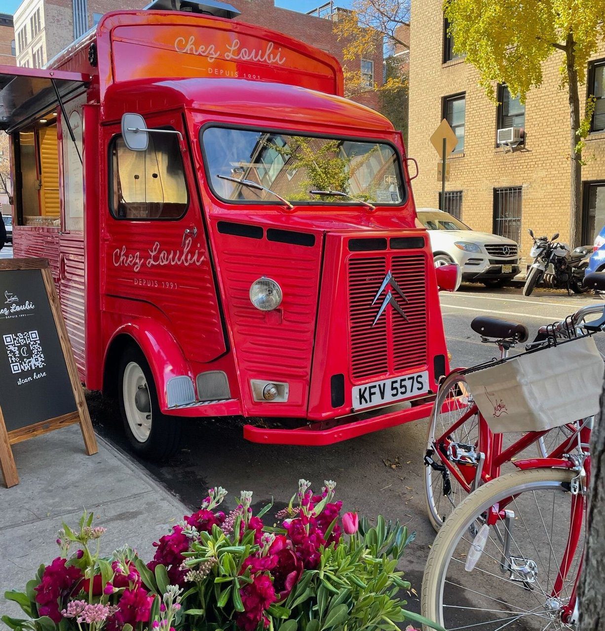Red vintage Citroën food truck labeled Chez Loubi parked by flowers with red bicycle in front.