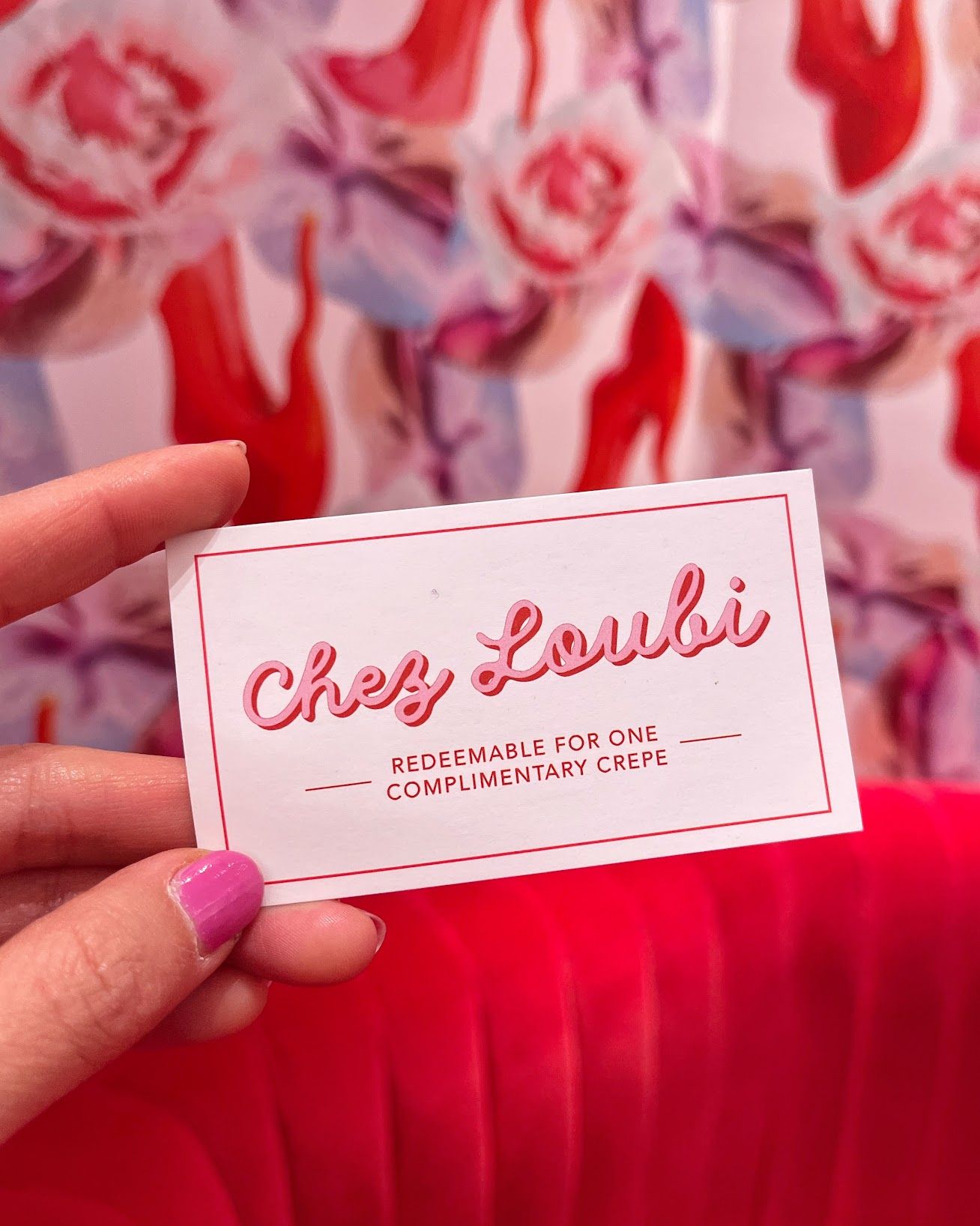 Hand with pink nails holds a “Chez Loubi” crepe card in front of a blurred floral background.