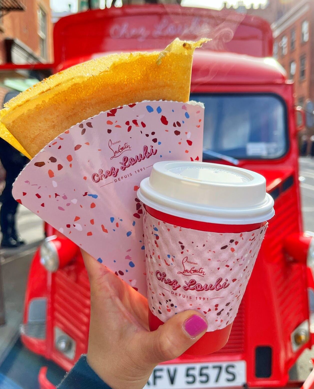 Hand holds a Chez Loubi crepe and coffee in front of a red vintage food truck on city street.