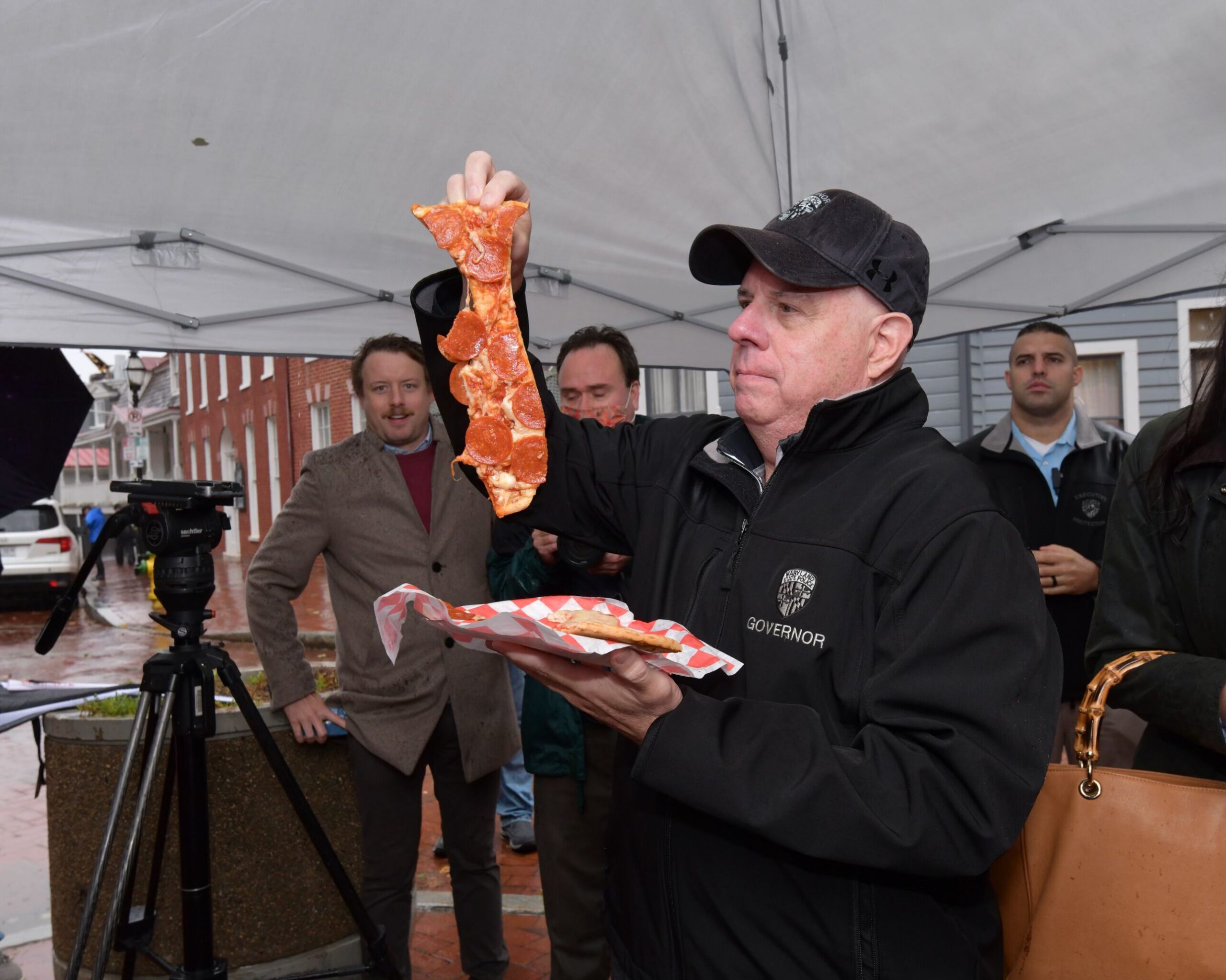 Man in black jacket holds a long slice of pepperoni pizza at an outdoor event; others watch.