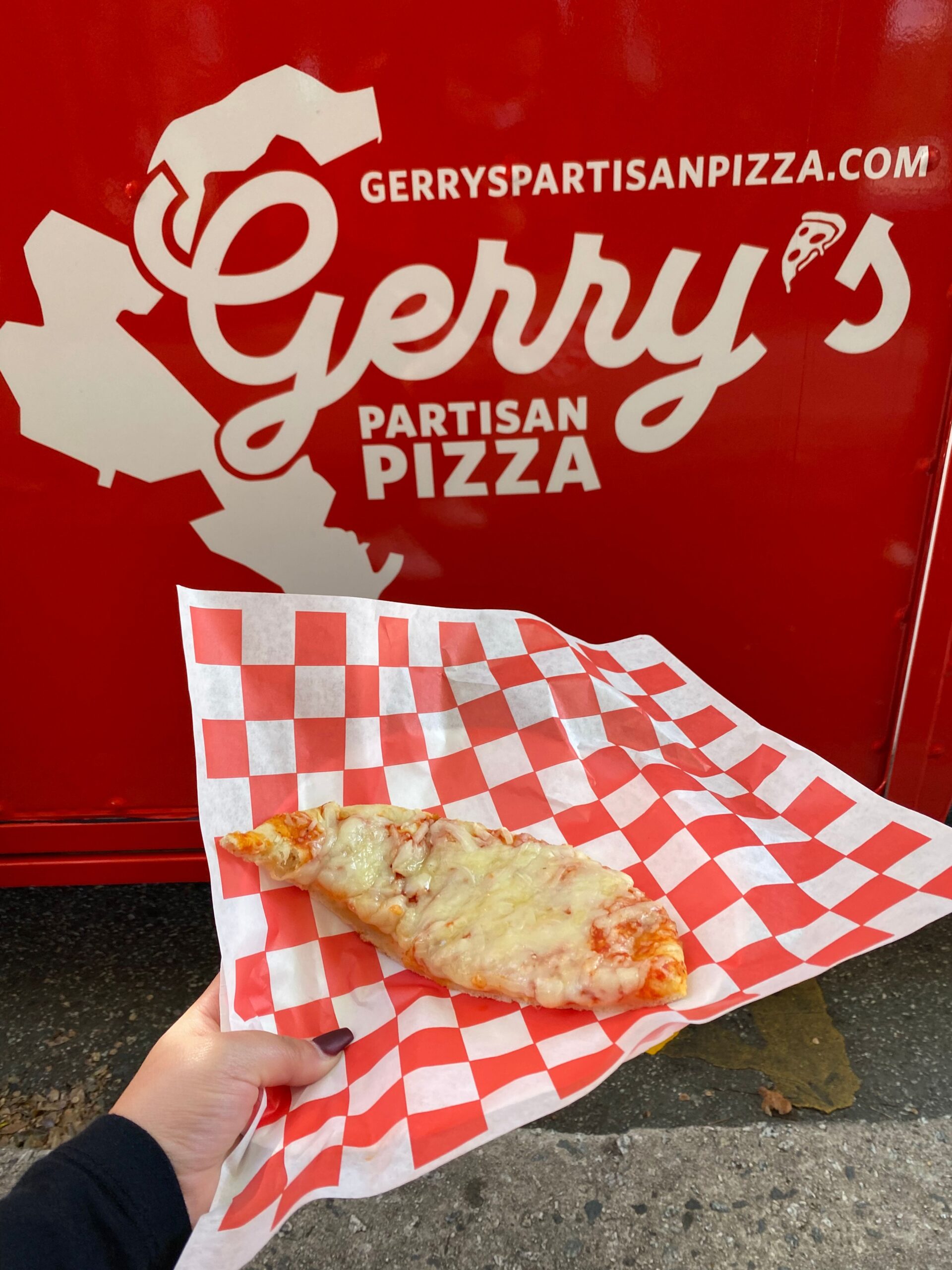 Hand holding a folded slice of pizza on checkered paper in front of Gerry’s Partisan Pizza truck.
